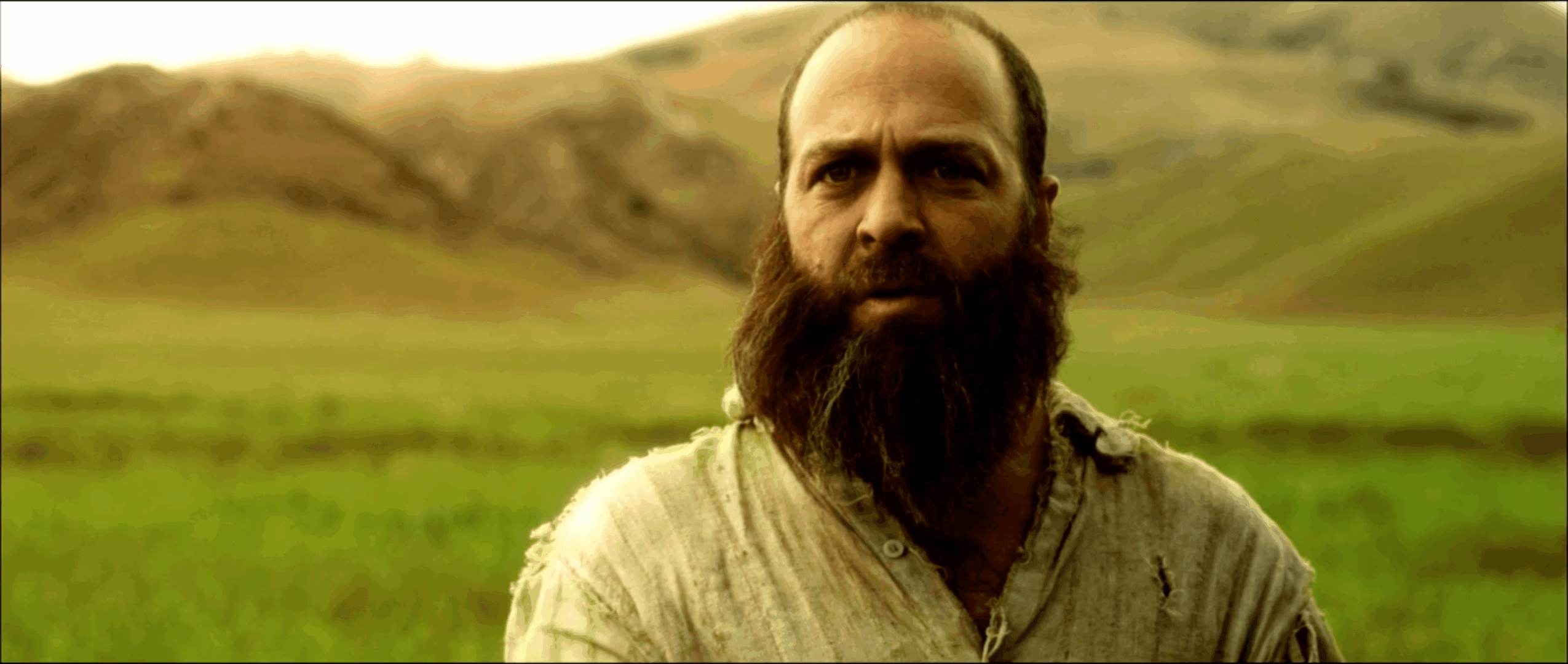Man with a beard and mustache in a grassy landscape with hills in the background.