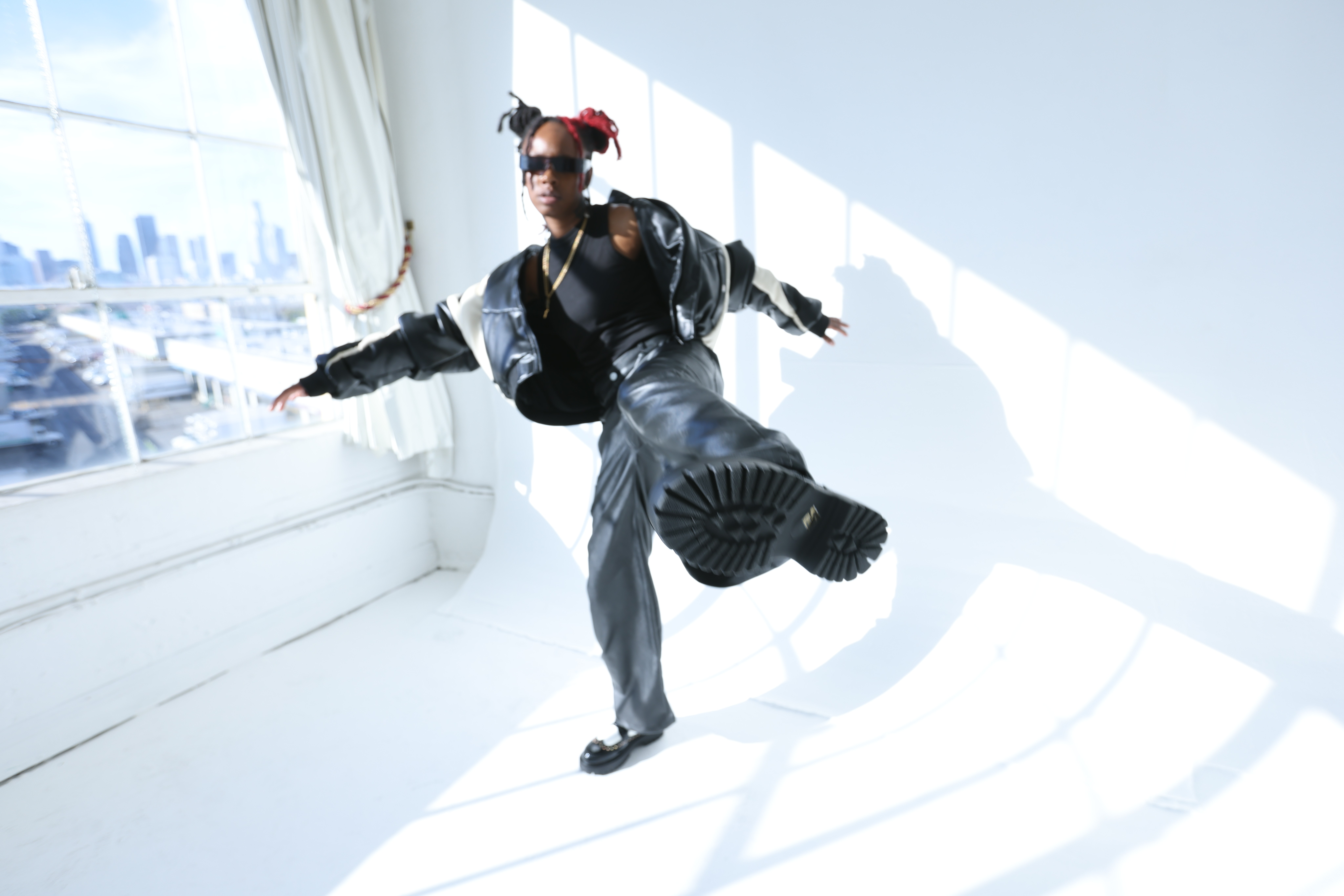 Person in a black jacket and gray pants balancing on one foot inside a bright room with large windows, city skyline outside
