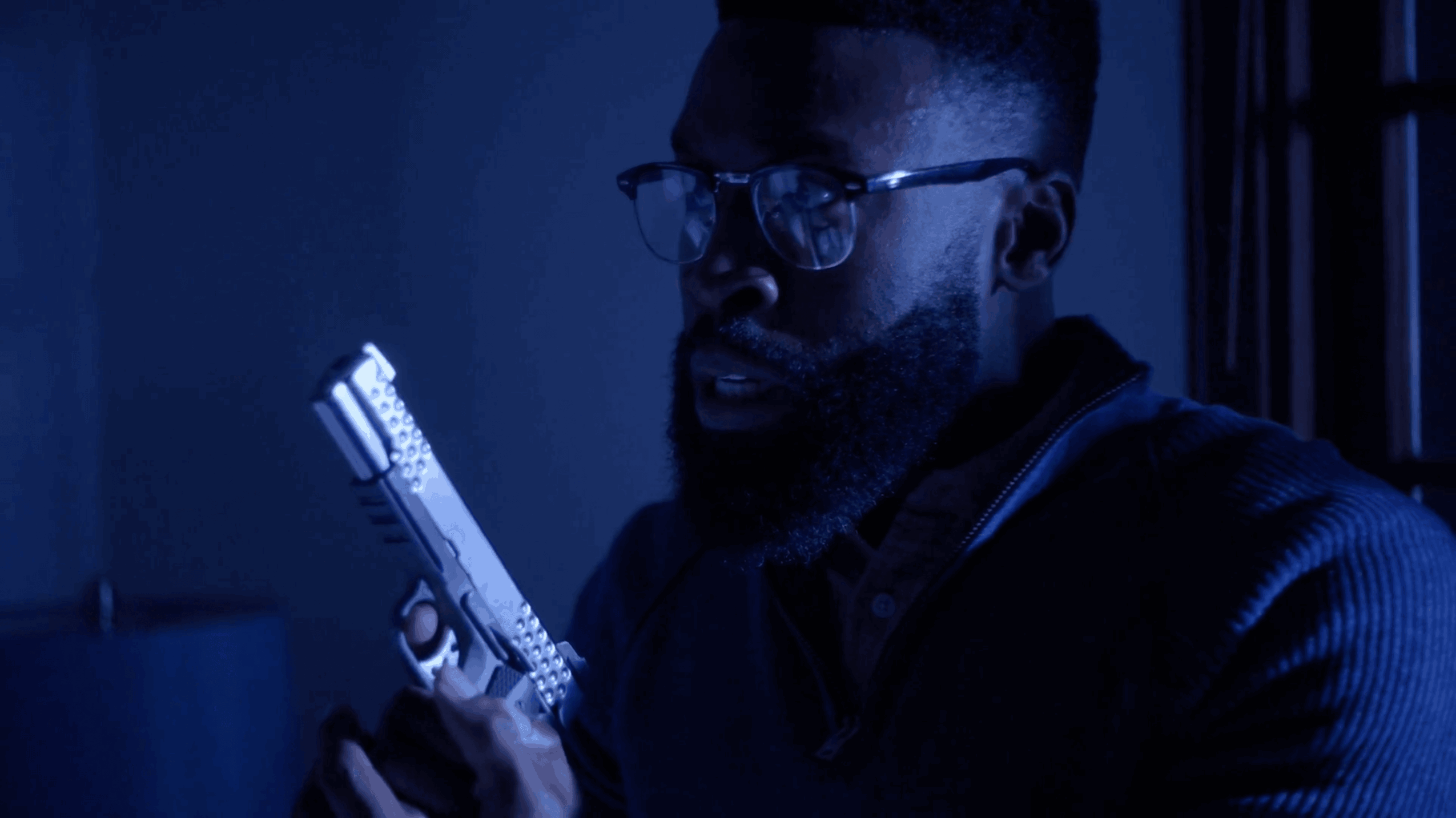 Man with glasses and beard holding a gun in a dimly lit room, looking to the side.