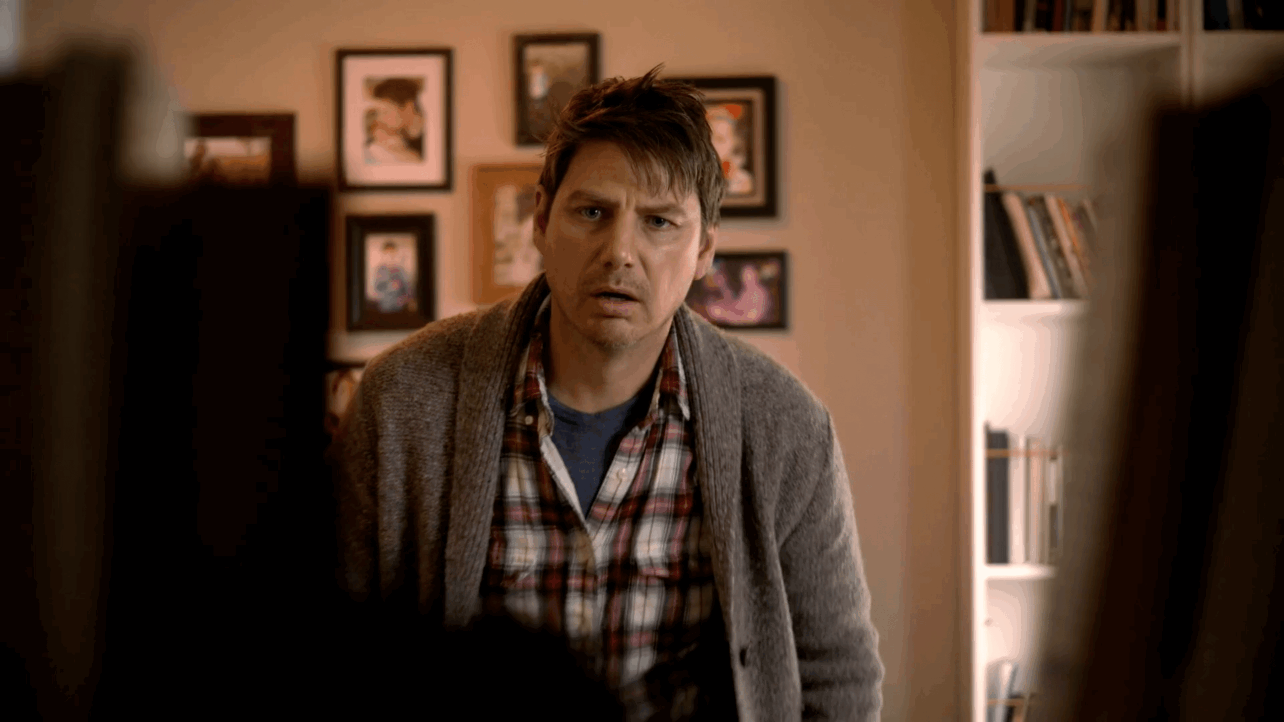 Man with dark hair and plaid shirt looks surprised indoors, with framed pictures on the wall behind him.