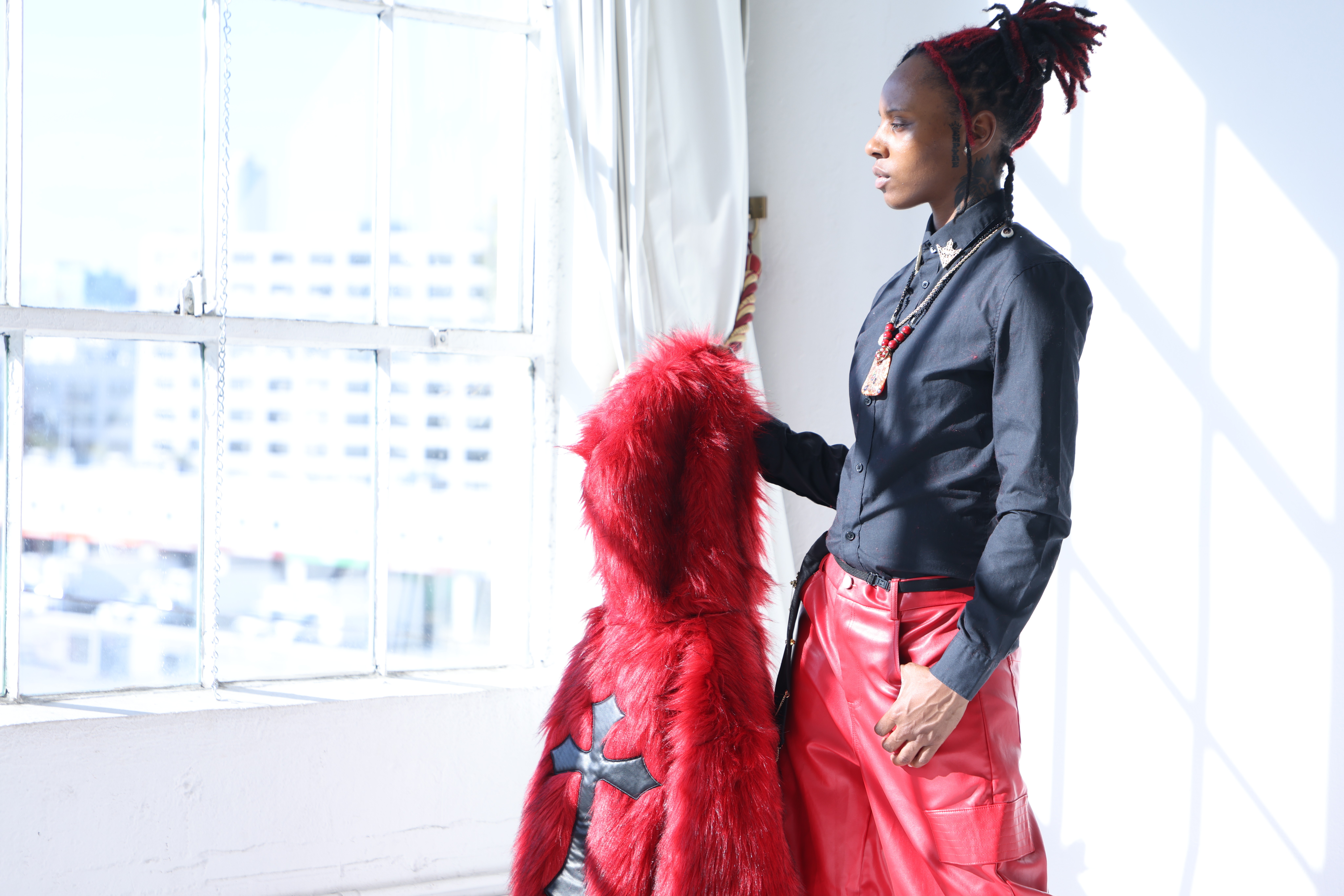 Woman with braided hair in a black jacket and red skirt stands near a window with city buildings outside.