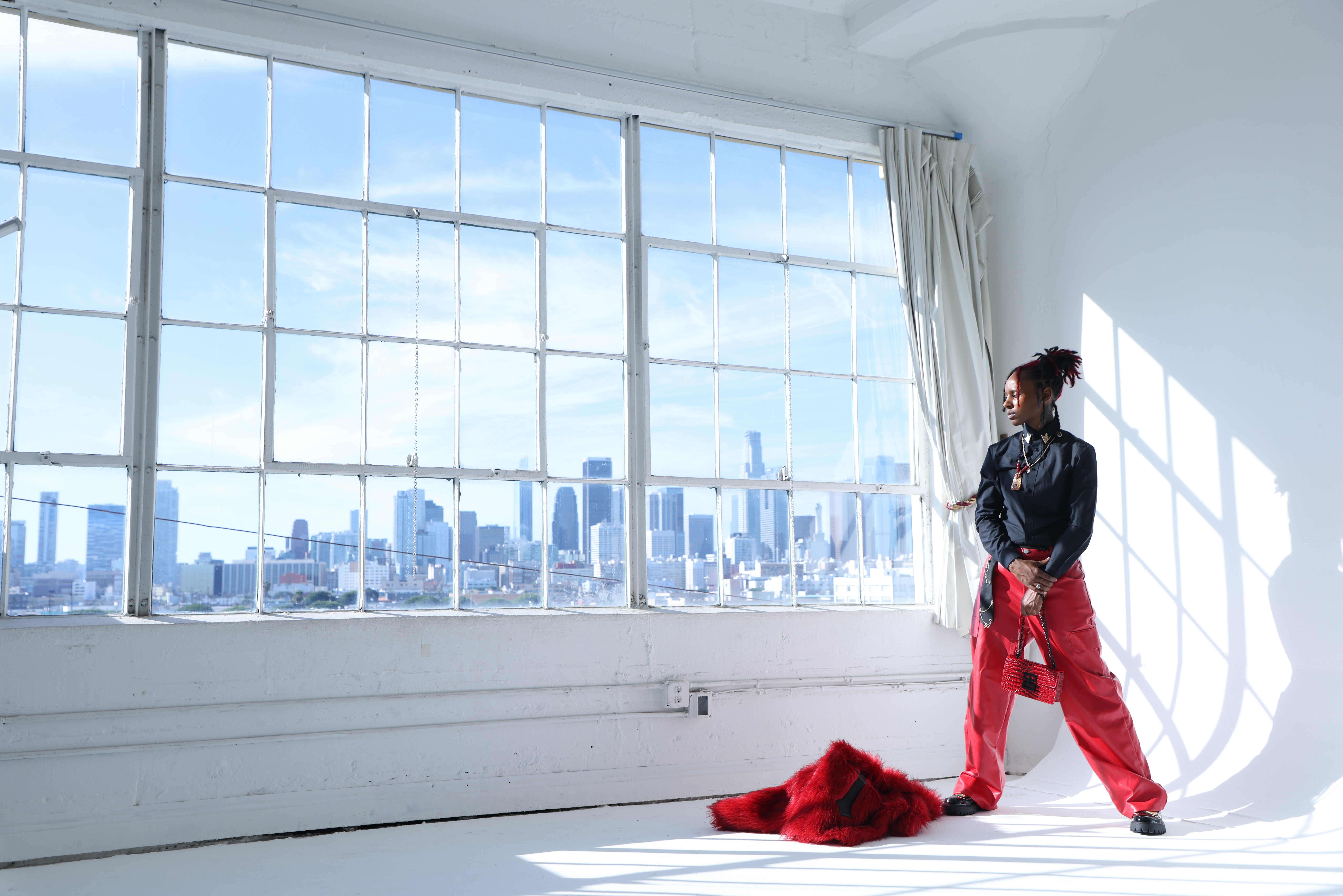 Woman standing near large window with city skyline view, sunlight casting shadows inside the room, white walls, and curtains.