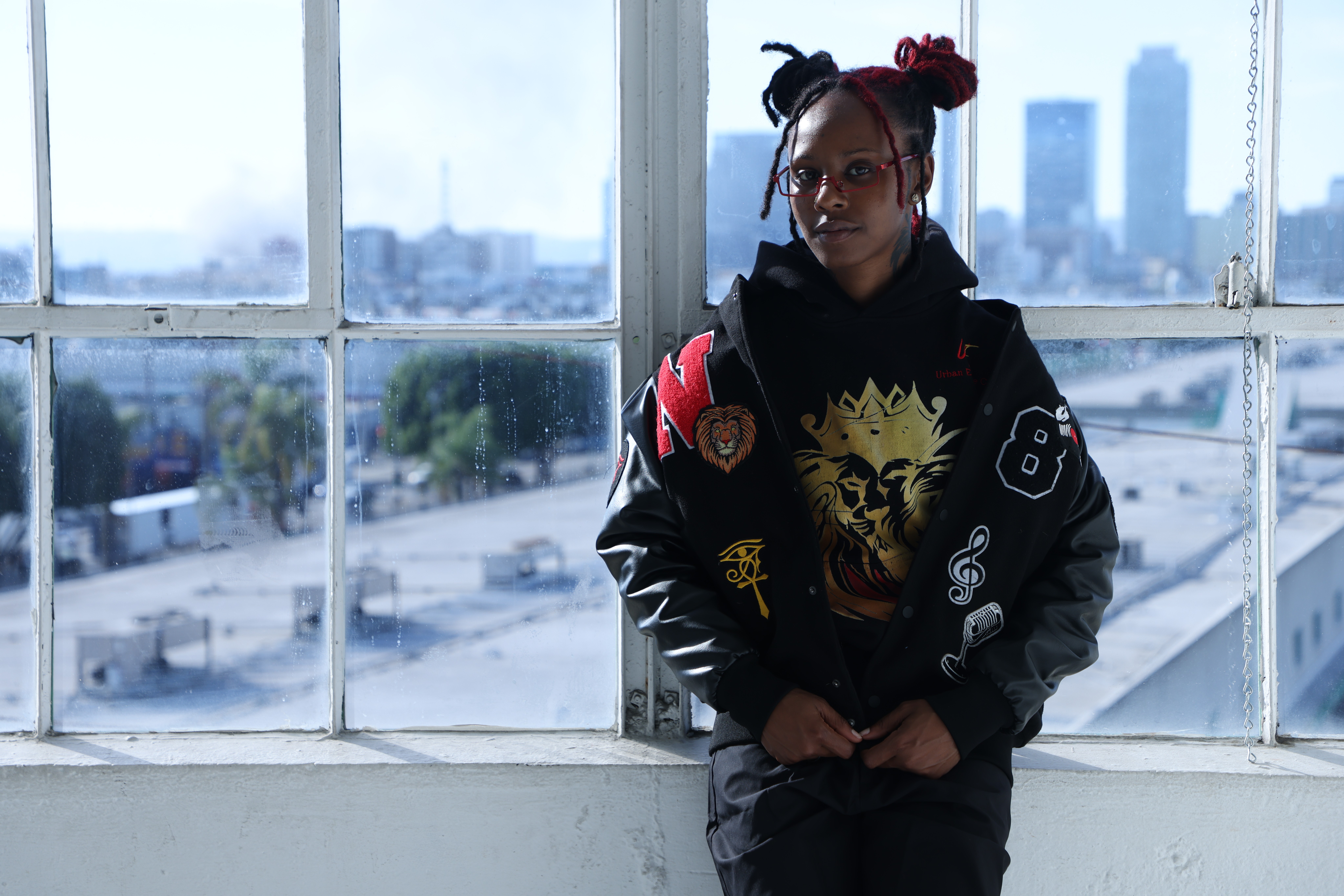 Person standing in front of large window with cityscape view, wearing a black jacket with colorful patches and a lion design, and glasses.
