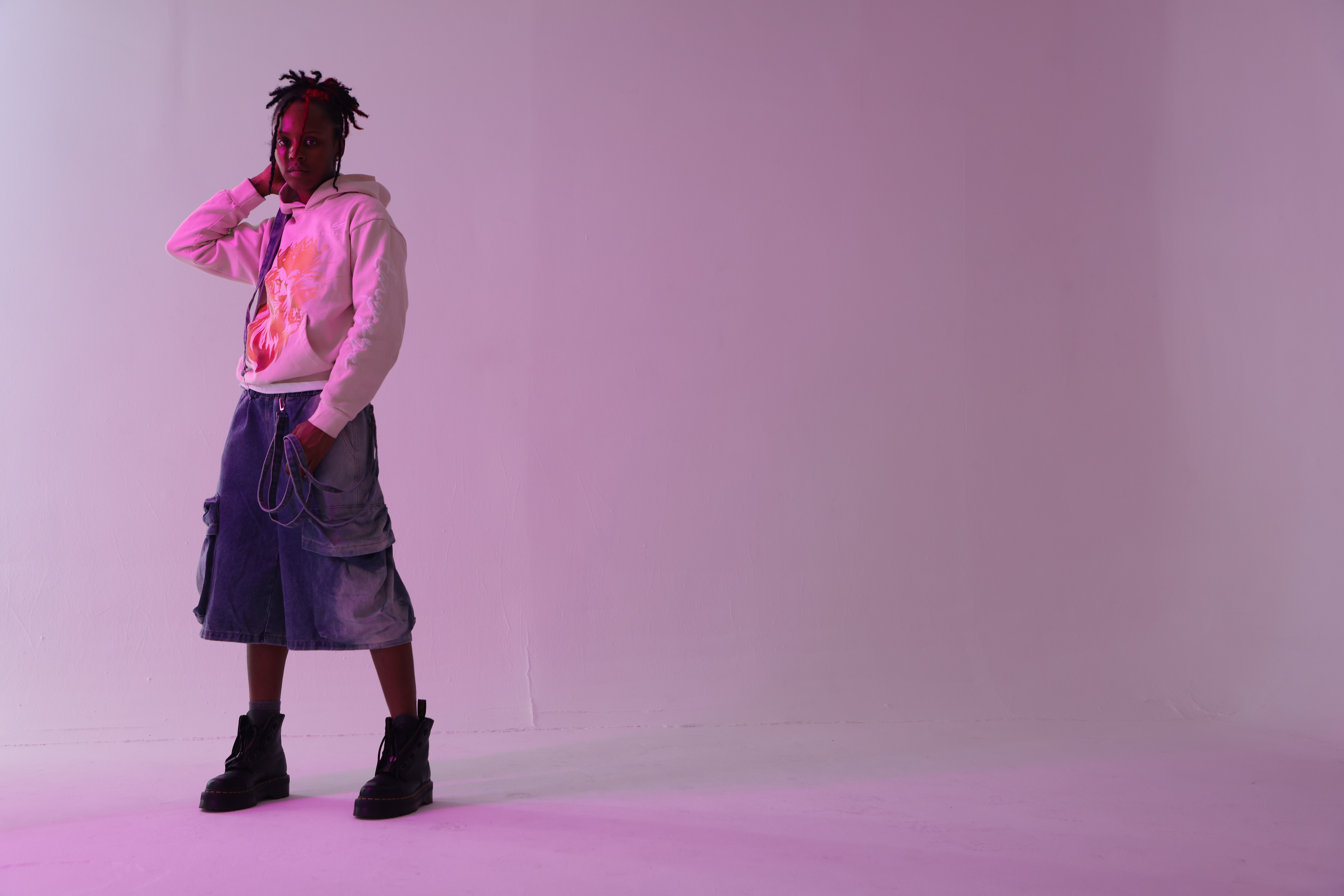 Person with dreadlocks wearing a hoodie, shorts, and boots standing against a plain background.