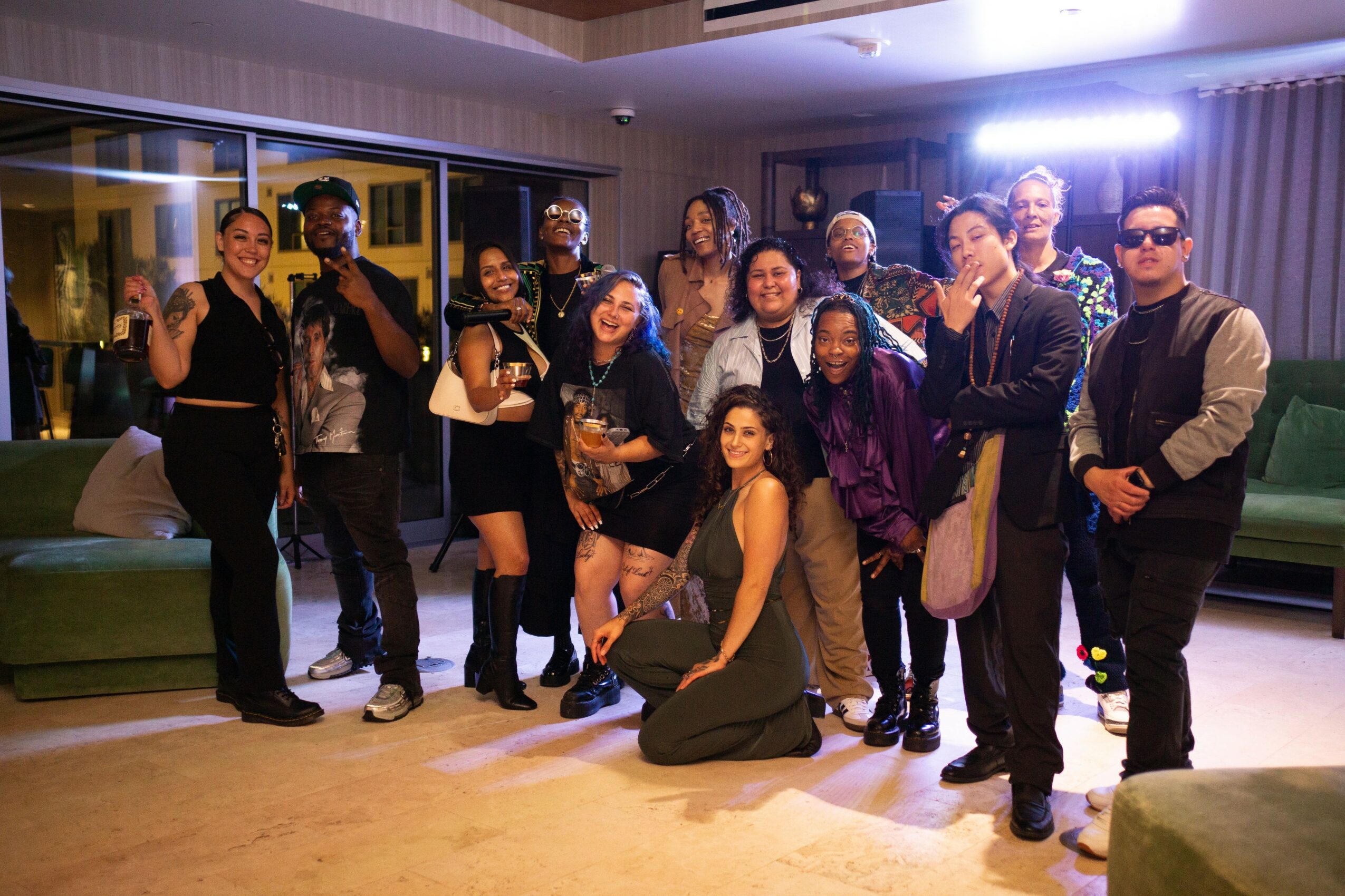 Group of 13 people posing indoors, some smiling and waving, with a window and curtains in the background.