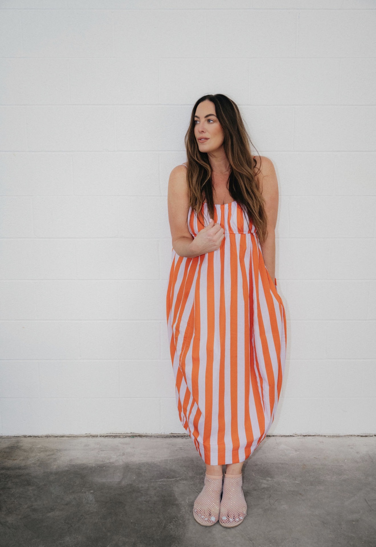 Woman in a striped orange and white dress standing against a white wall, looking to the side.