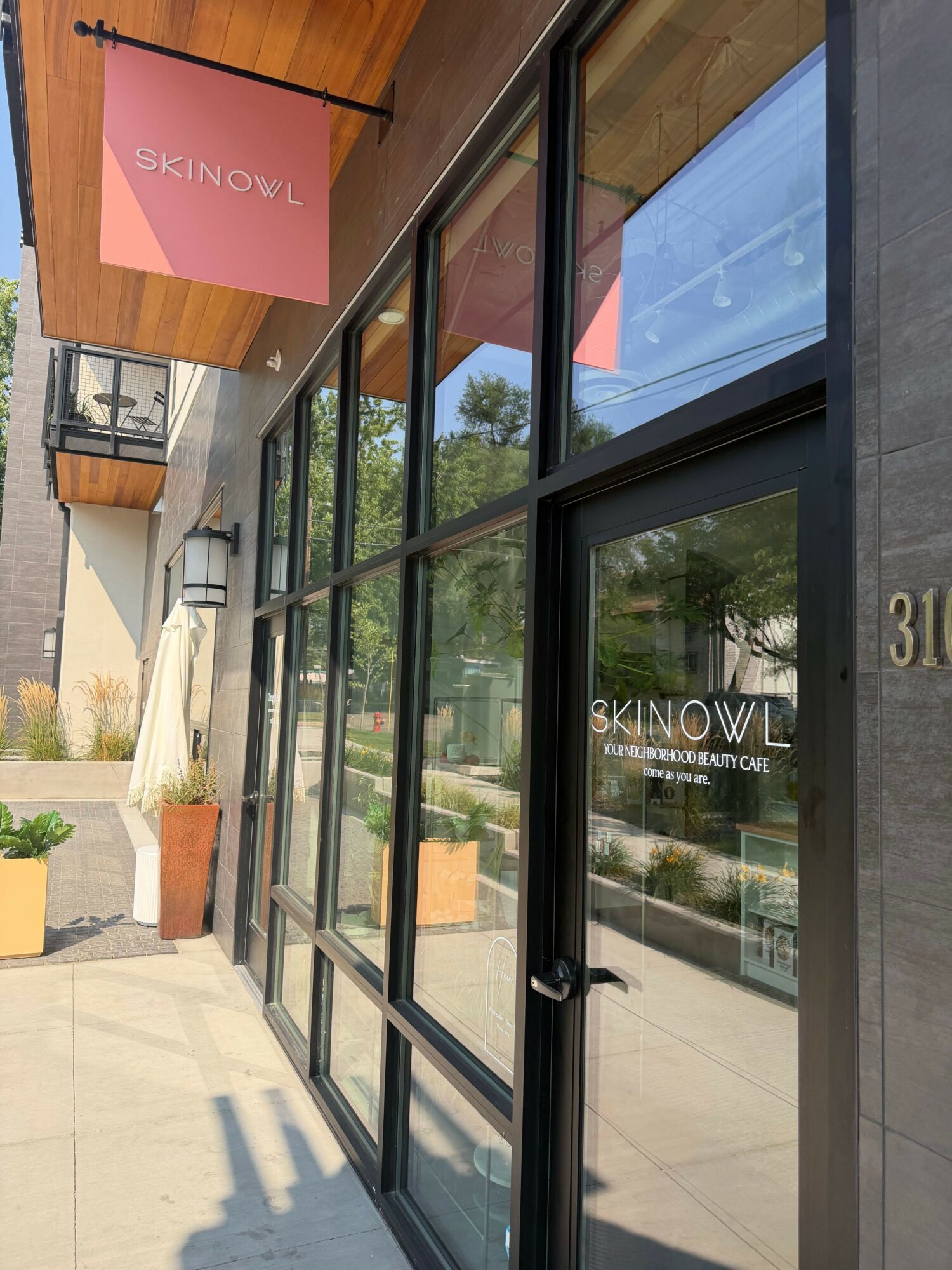 Glass door with reflections of trees and sky, with a pink sign reading 'SKINOVIA' nearby, and a sidewalk outside.