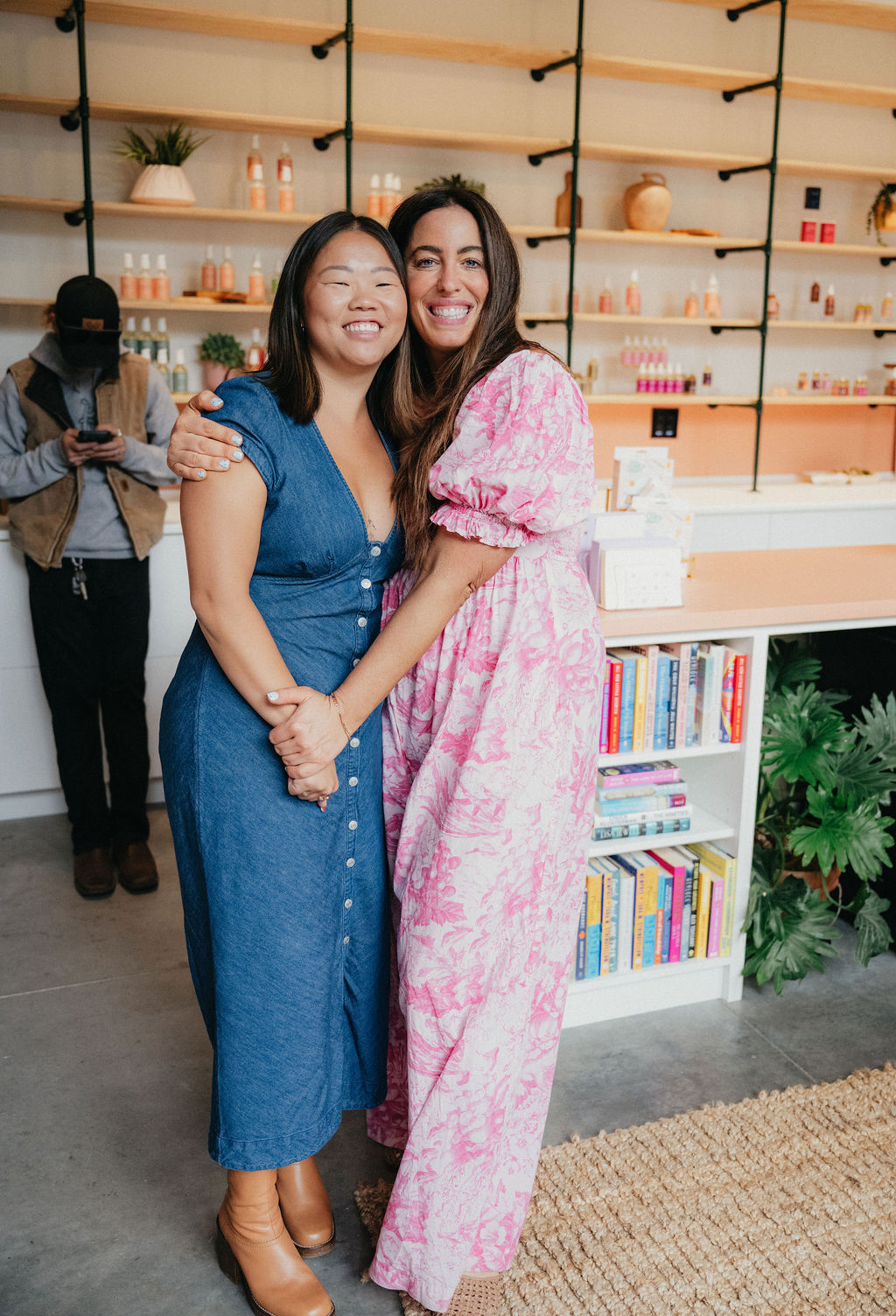 Two women smiling and hugging in a store with shelves of products and books behind them.