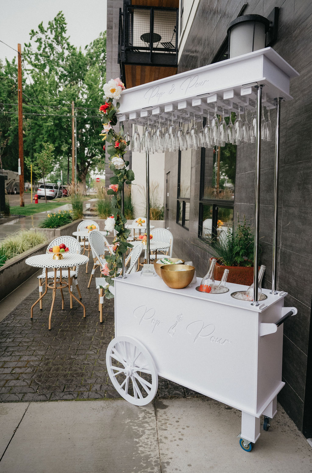 White mobile bar with glasses and flowers outside a building, set for an event with tables and chairs nearby.