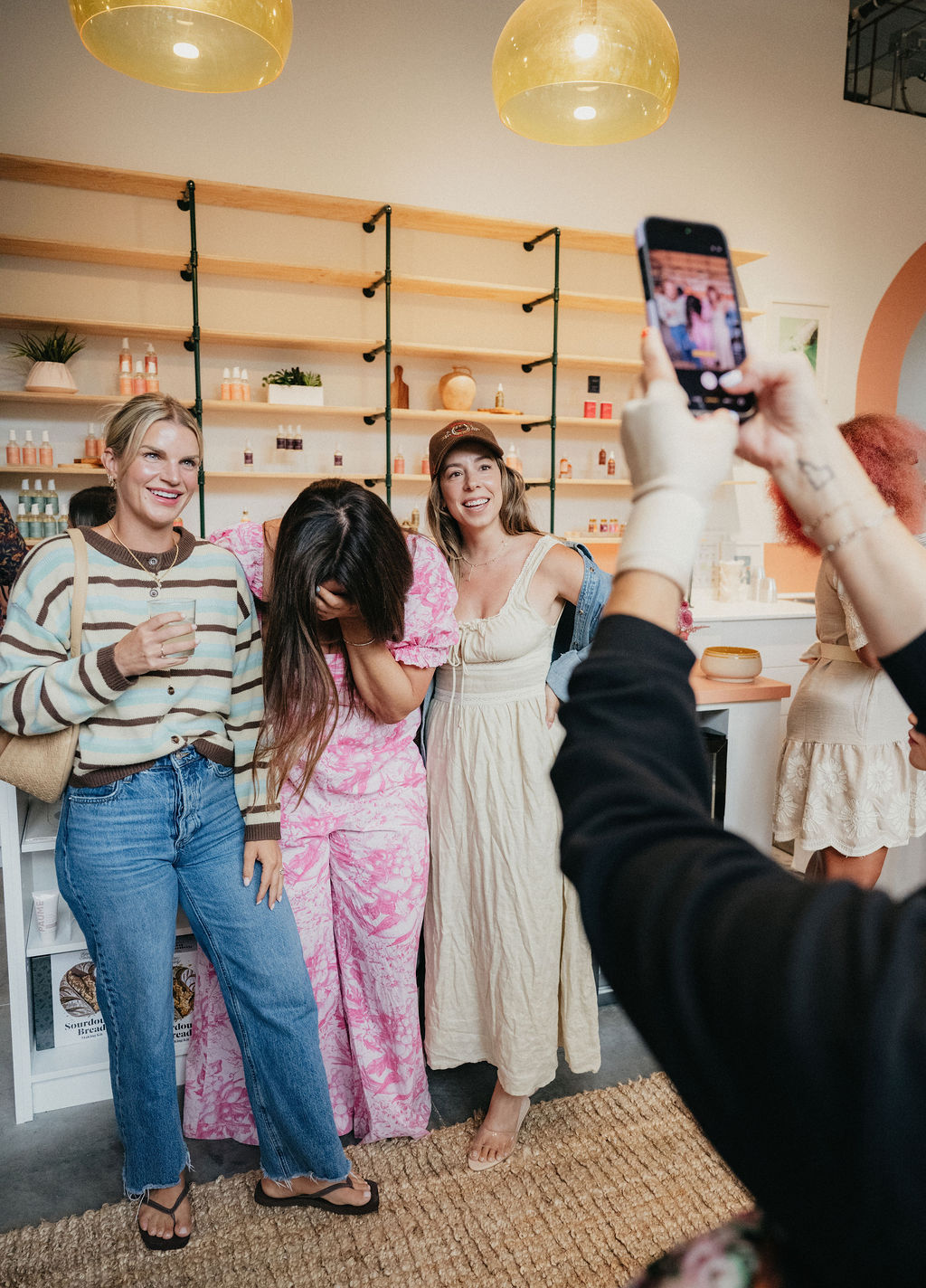 Three women stand together smiling, one taking a photo with a smartphone in a cozy, decorated room.