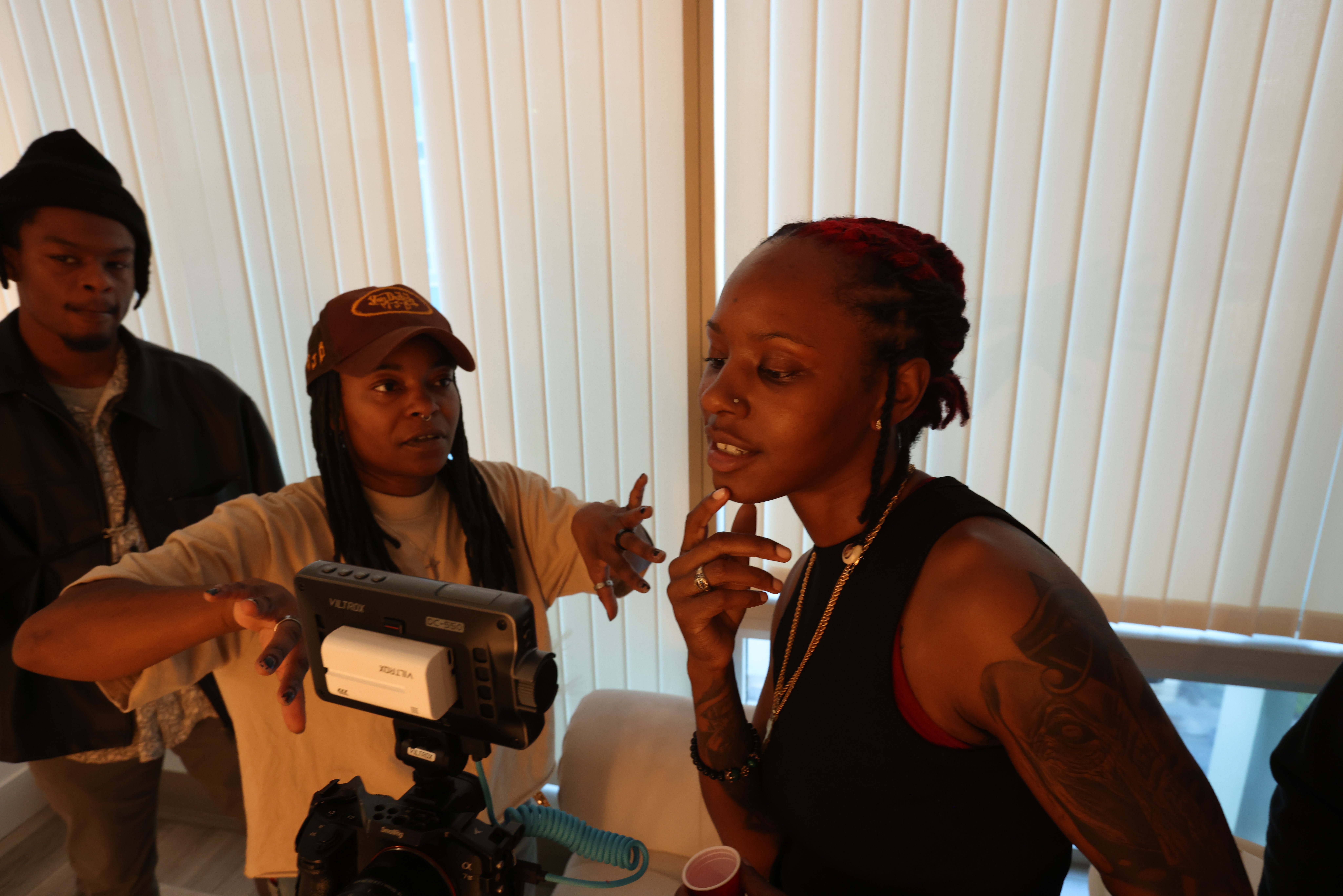 Three women engaged in conversation indoors near window blinds, with one woman holding a camera or recording device, in a casual setting.