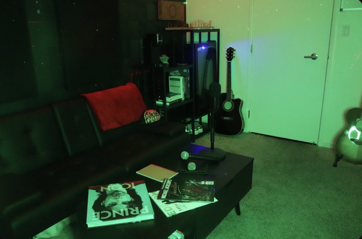 Living room with black sofa, red blanket, coffee table with magazines, guitar, and shelves with electronic equipment, illuminated by green and blue lights.