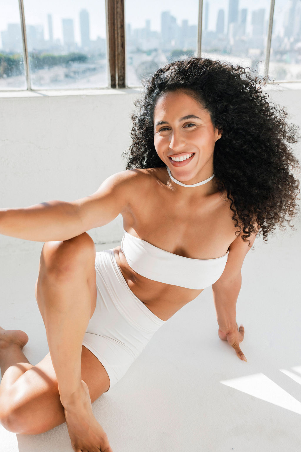 Smiling woman with curly hair in white outfit sitting on floor near window, city skyline in background.
