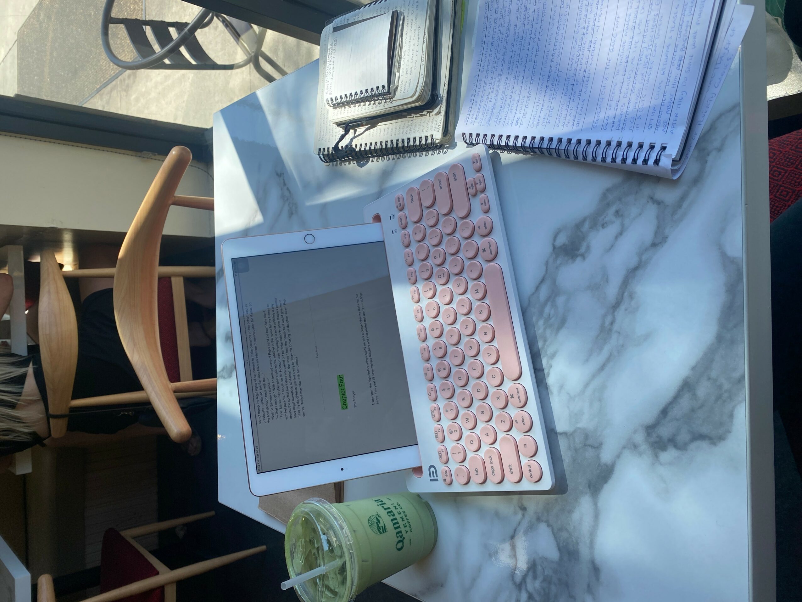 Tablet, keyboard, notebooks, and a drink on a marble table, sunlight streaming in, in a bright room.
