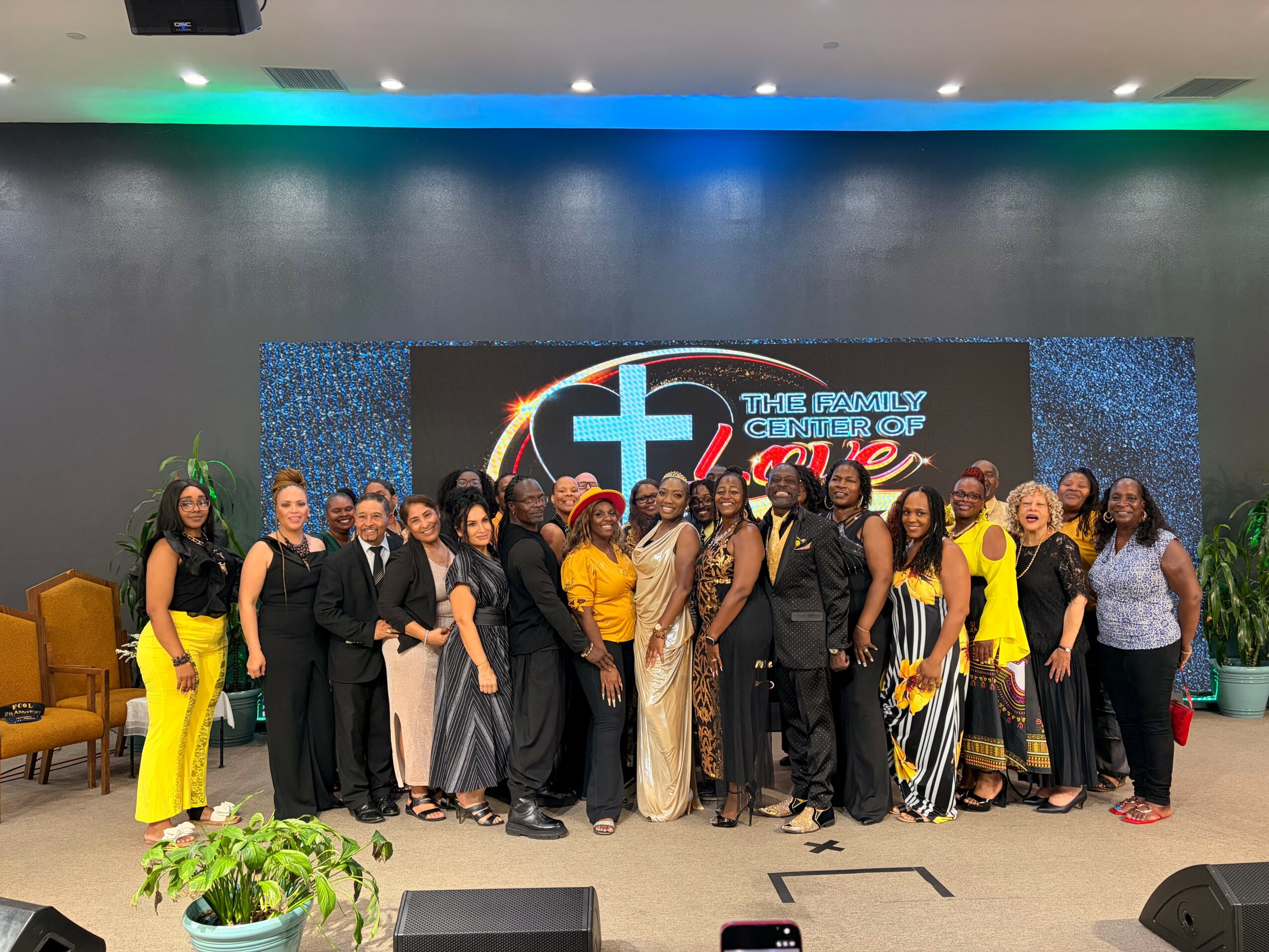 Group of people standing on stage in front of a large screen with a logo and text, some wearing formal attire, others in casual clothing.