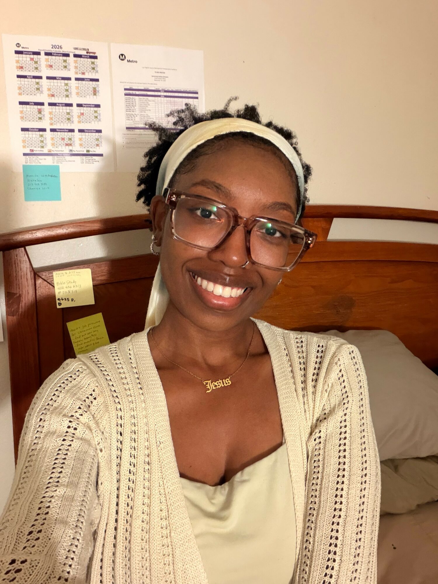 Smiling woman with glasses, wearing a headband and cream-colored cardigan, sitting on a bed in a room with calendar and notes on wall.