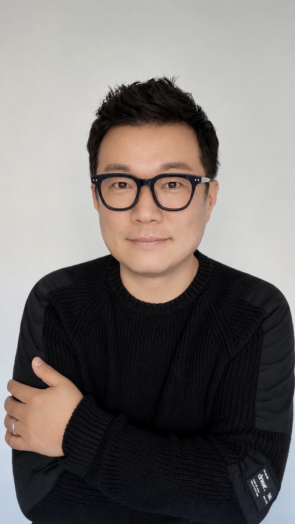 Portrait of a person with short dark hair, glasses, wearing a black sweater, smiling slightly, arms crossed, against a plain background.