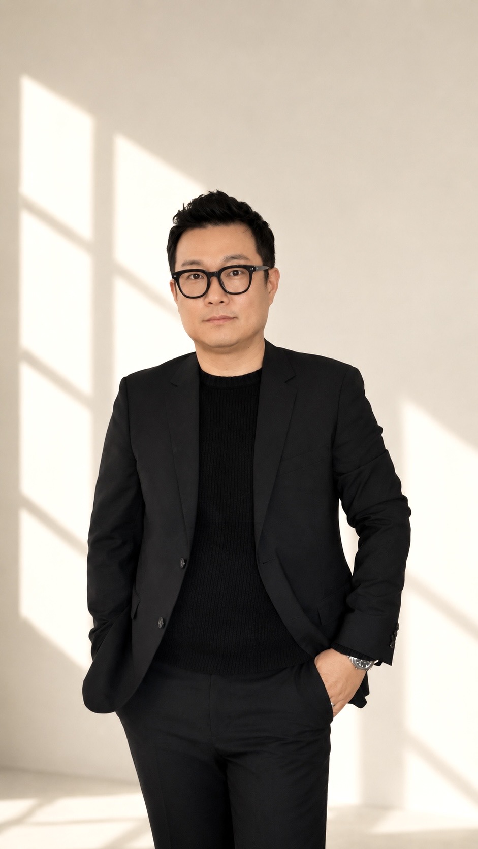Man in black suit and glasses standing with hands in pockets against a light background with window shadows.