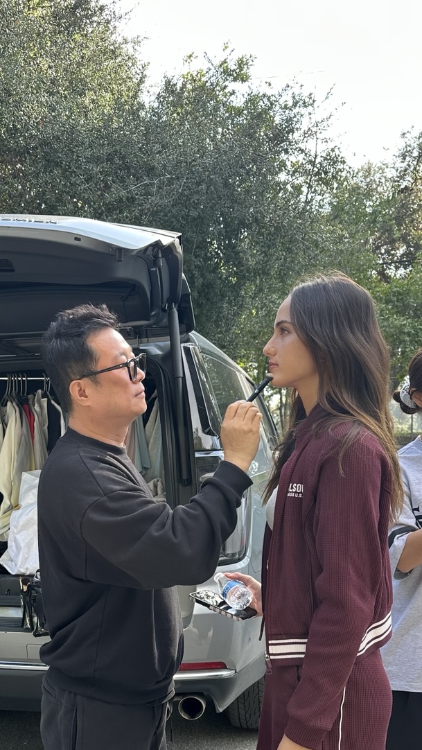 A person with glasses applies makeup or face paint to a young woman outdoors near a vehicle, with trees in the background.
