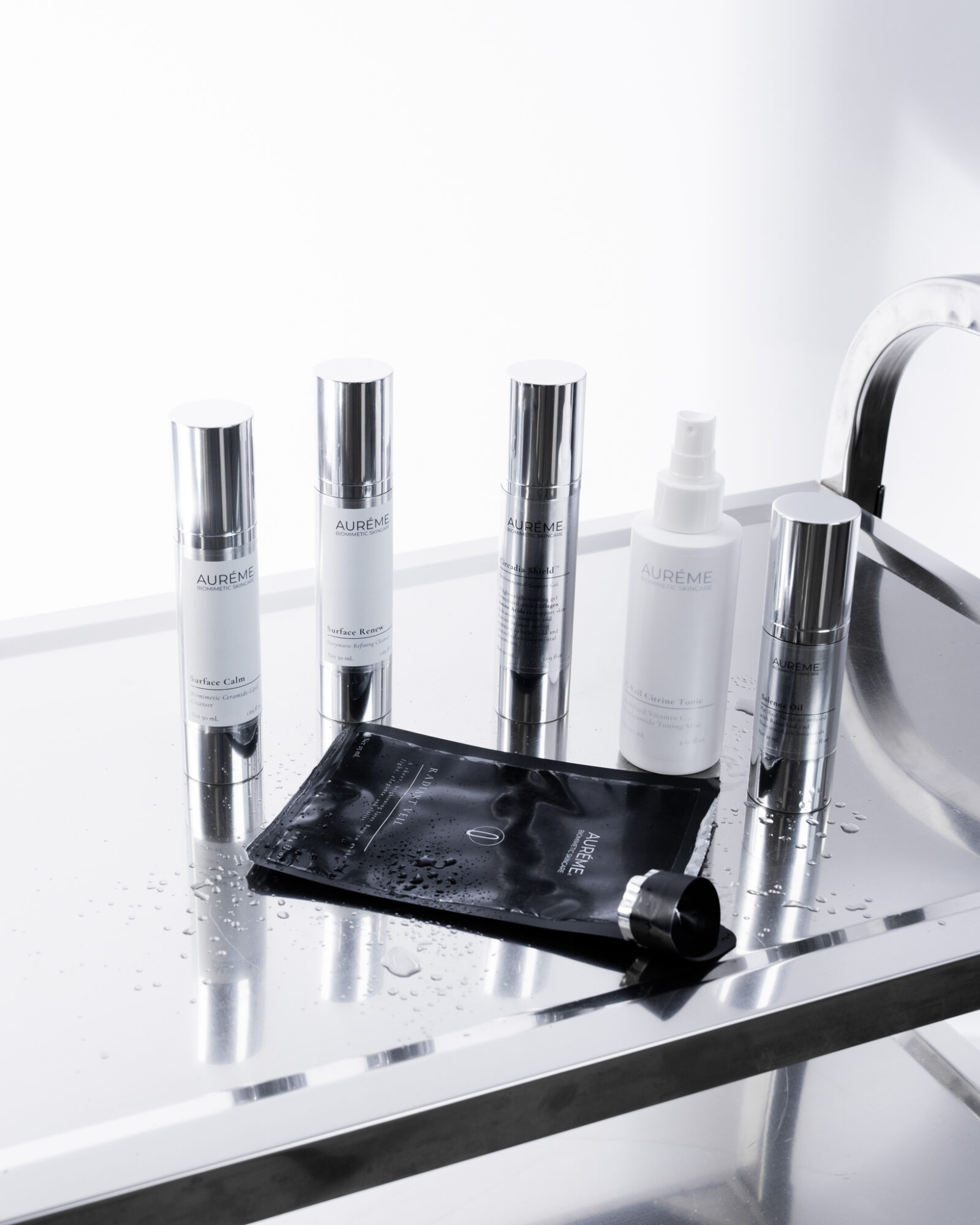 Skincare products and a black applicator on a reflective surface with a white background.