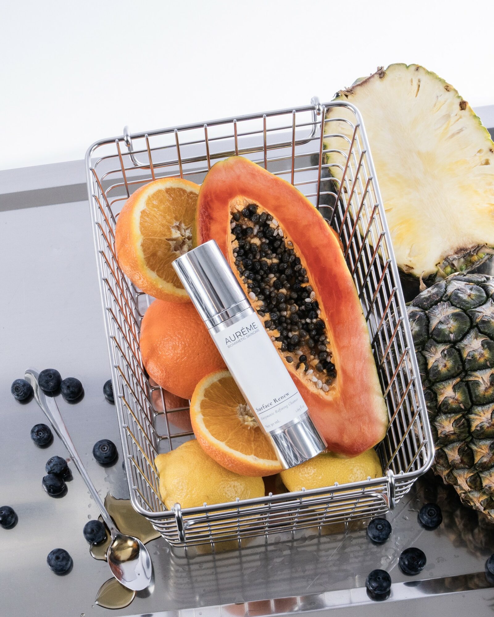 A small metal basket filled with sliced papaya, oranges, and lemon, with a tube of skincare product on top, on a white surface with scattered blueberries and a pineapple in the background.