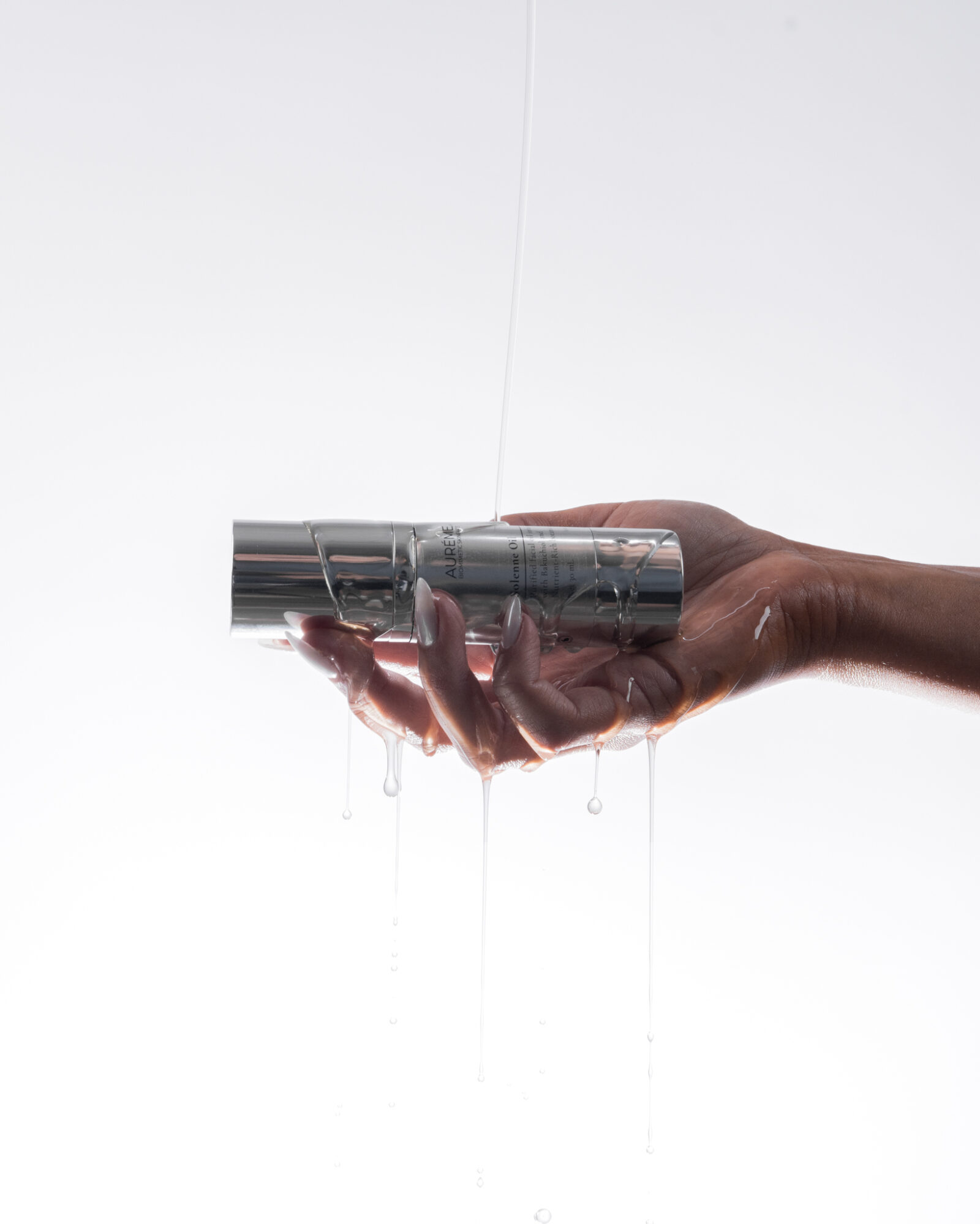 Hand holding a metallic object under running water, with water dripping down, against a plain white background.