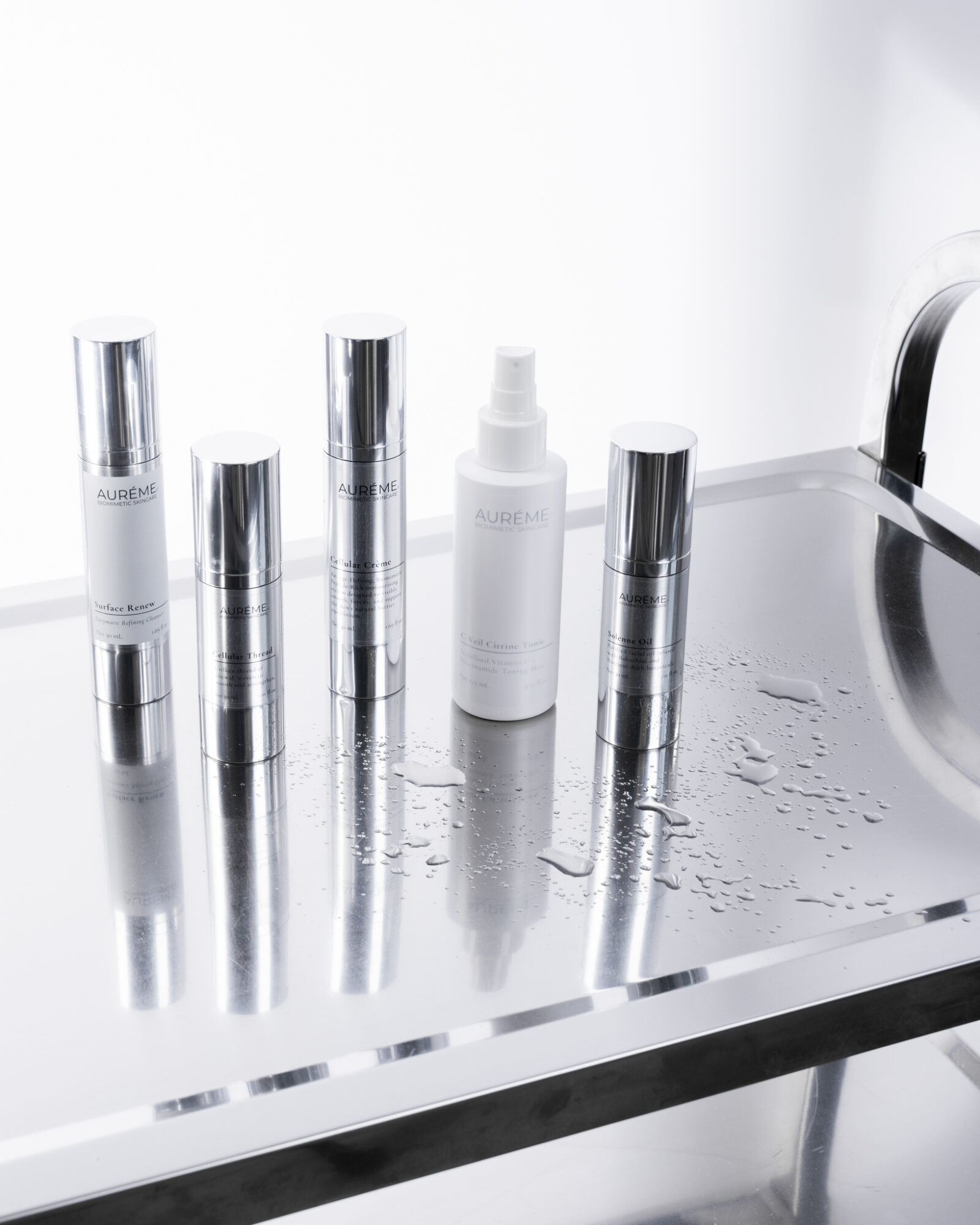 Five silver cylindrical bottles and one white spray bottle on a wet stainless steel surface, with a sink faucet visible on the right.