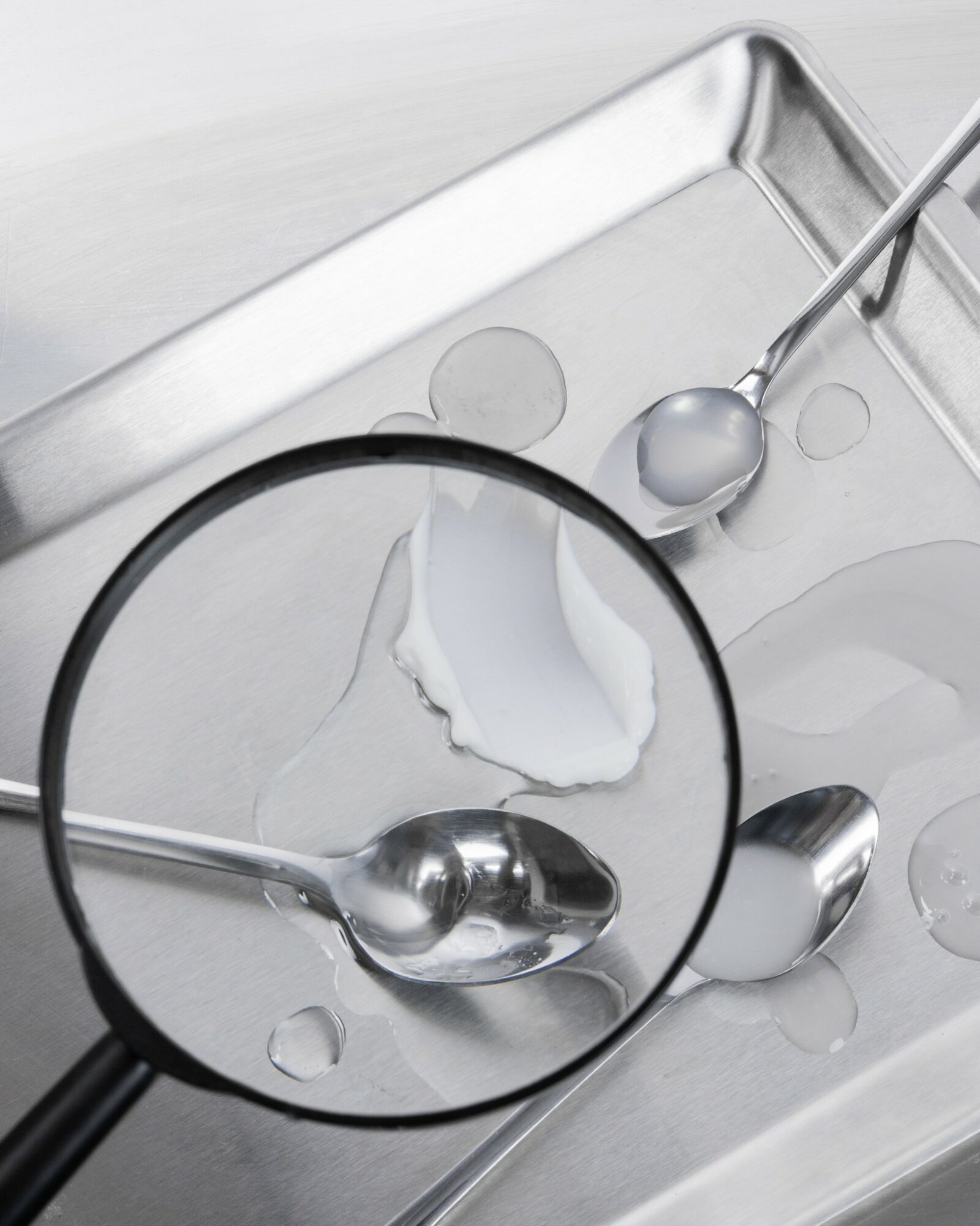 Stainless steel sink with a spoon and a ladle, magnified by a magnifying glass, with water droplets visible.
