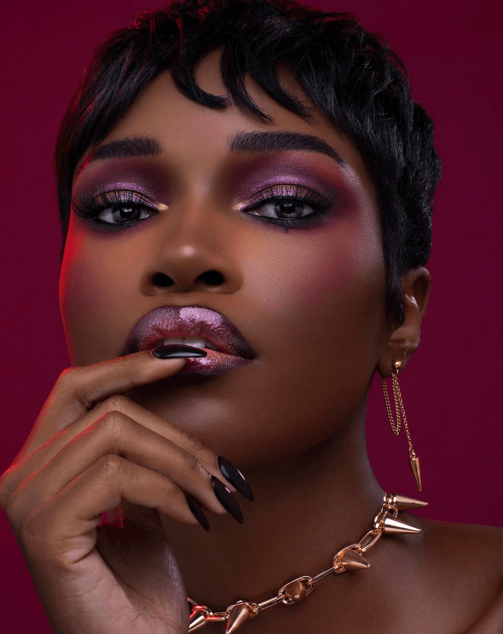 Close-up of a woman with short black hair, purple eye makeup, and gold jewelry, touching her lips.