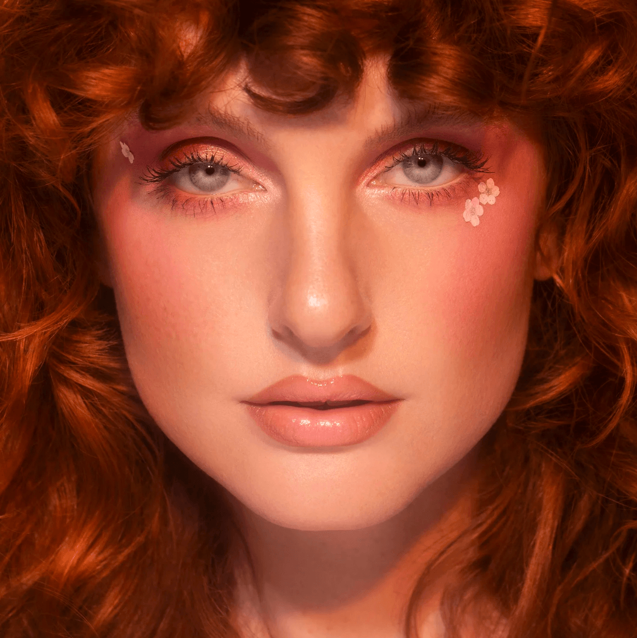 Close-up of a woman with curly red hair, pink eye makeup, and floral face decorations, looking directly at the camera.