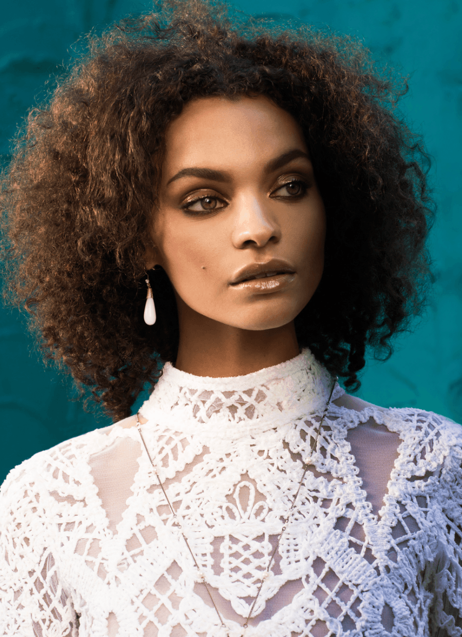 Portrait of a woman with curly hair wearing a white lace top and earring, against a teal background.