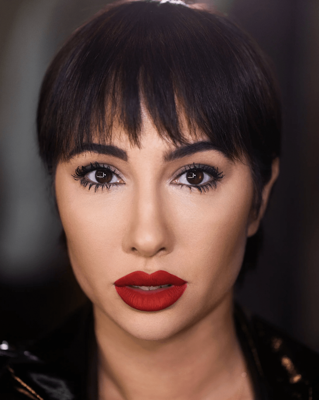 Close-up of a woman with short dark hair, bold makeup, and red lipstick, looking directly at the camera.