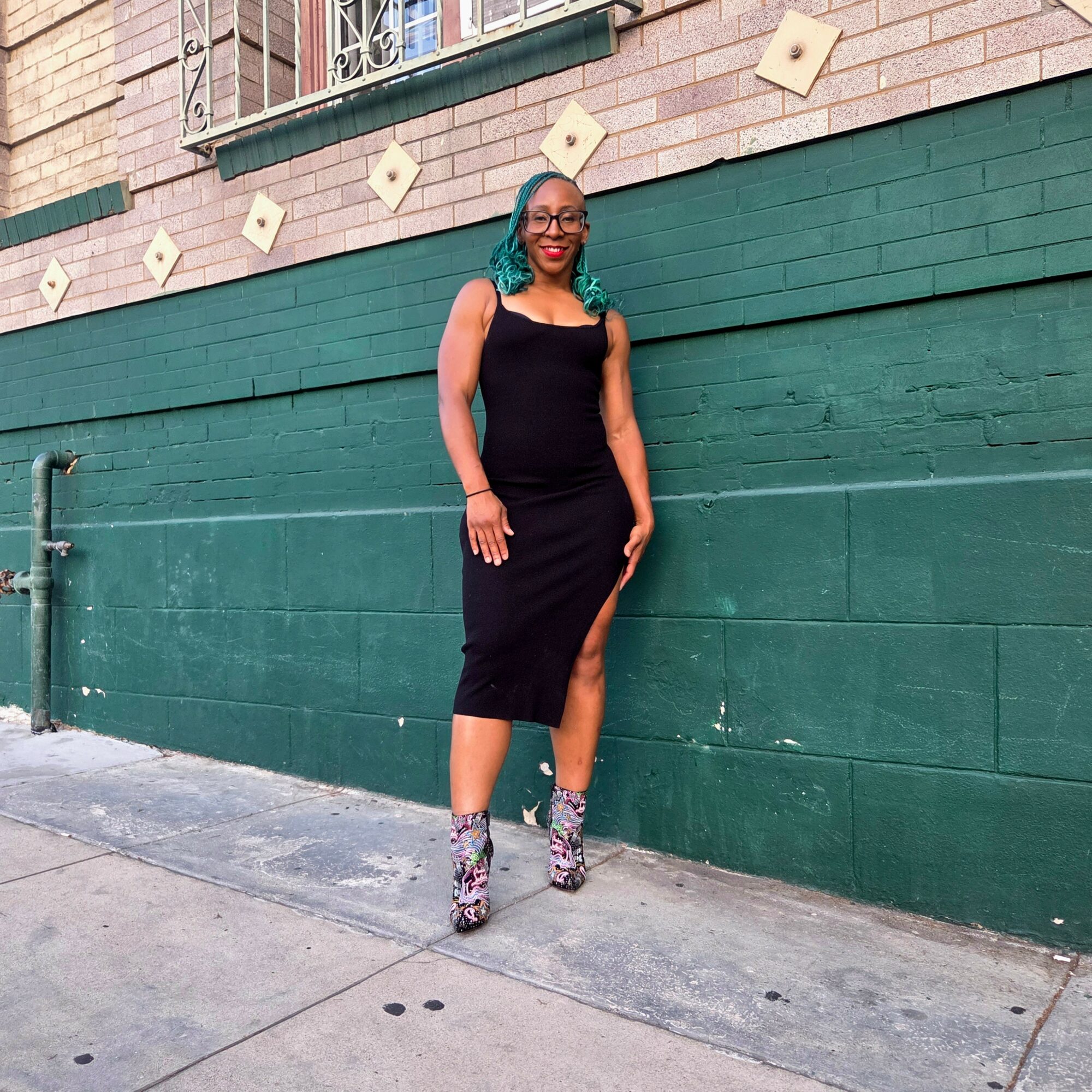 Woman with teal hair in a black dress and patterned boots standing against a green wall outdoors.