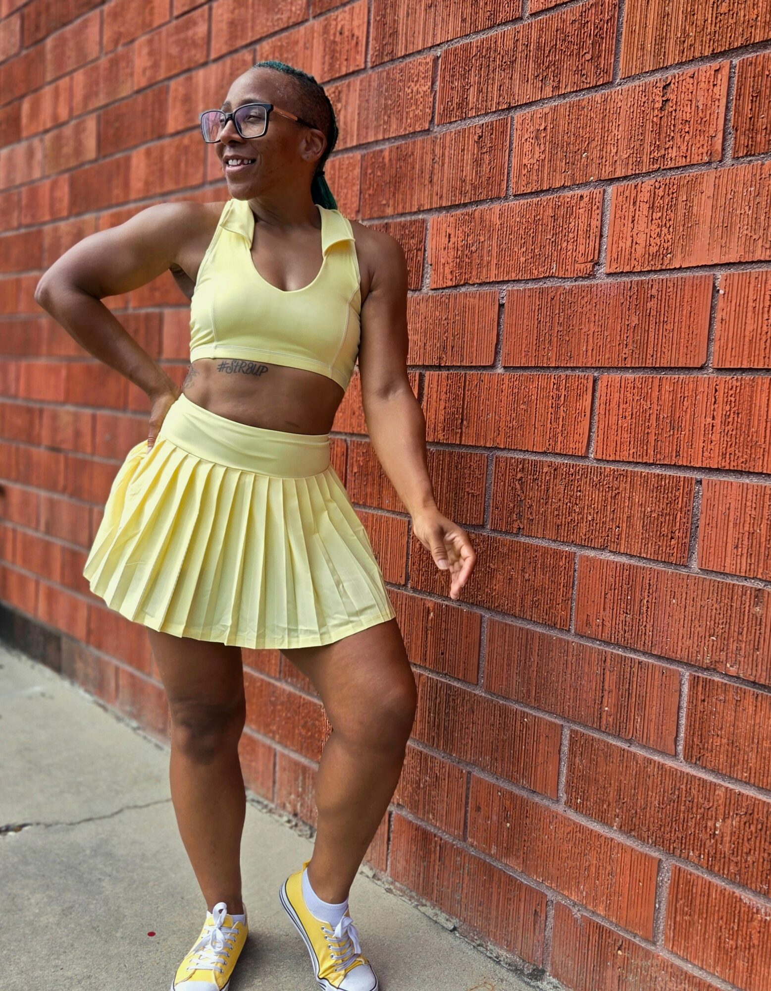 Woman in yellow sports bra and pleated skirt standing against a brick wall, smiling, with one hand on her hip.