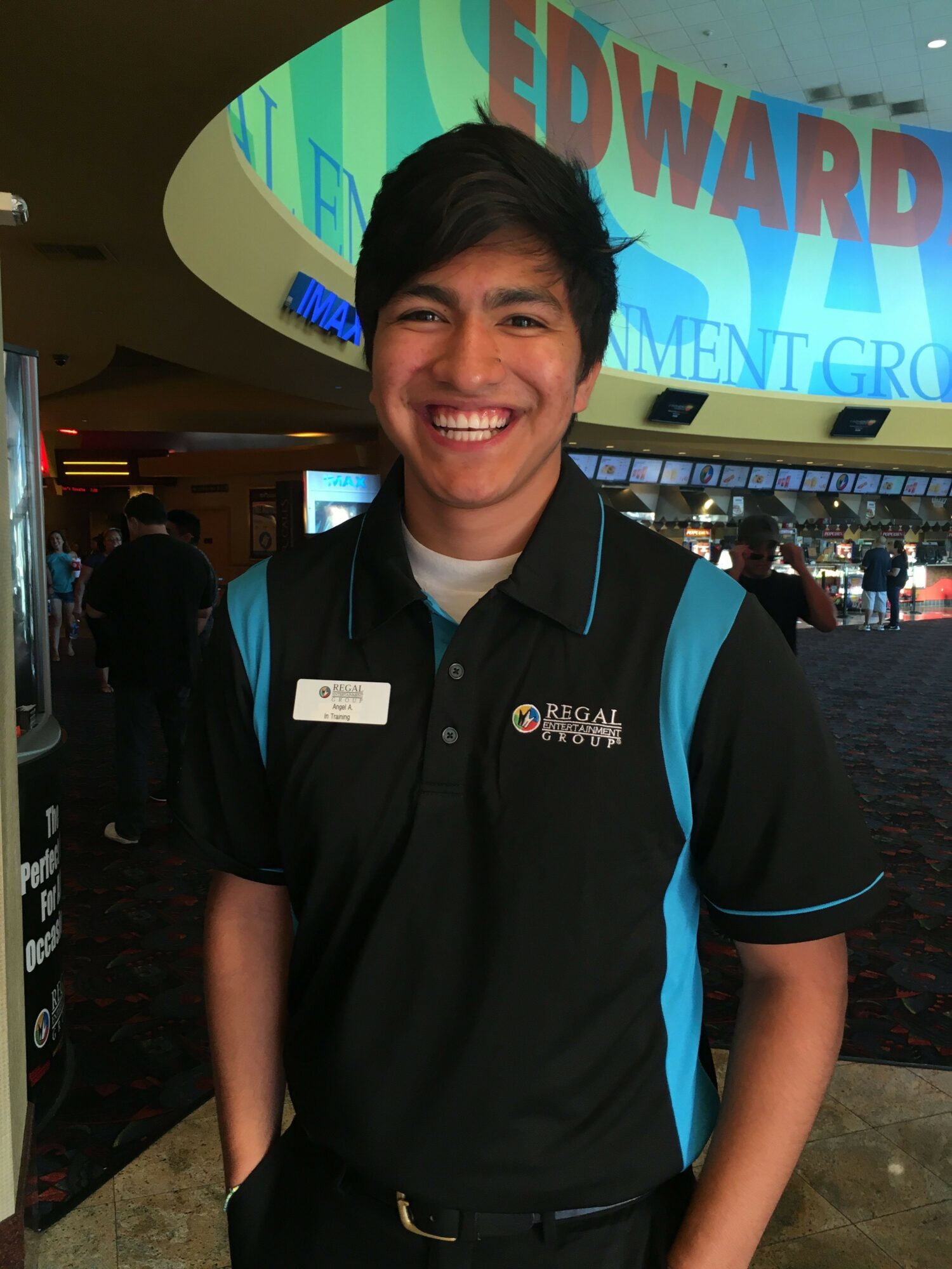 Smiling person in black and blue uniform standing indoors with a large screen behind them.