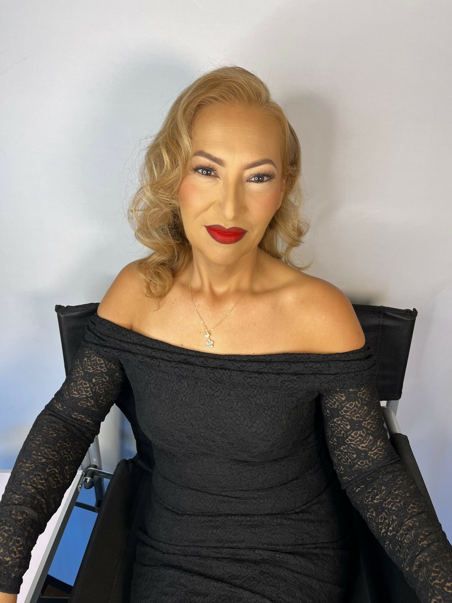 Woman with blonde hair, red lipstick, wearing an off-shoulder black dress, sitting against a white background.