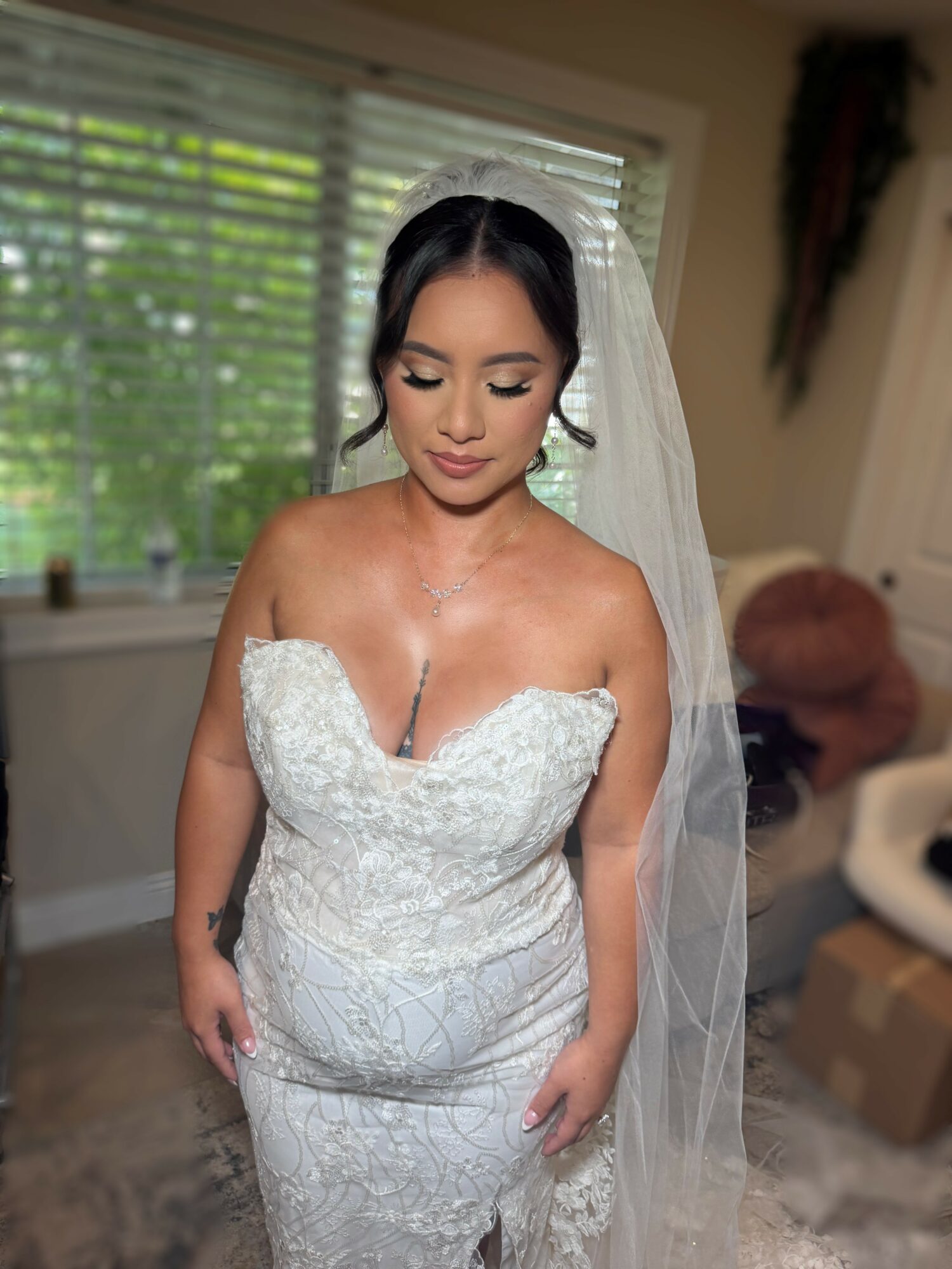 Woman in wedding dress with veil, standing indoors near window with blinds, looking down, with a blurred background.