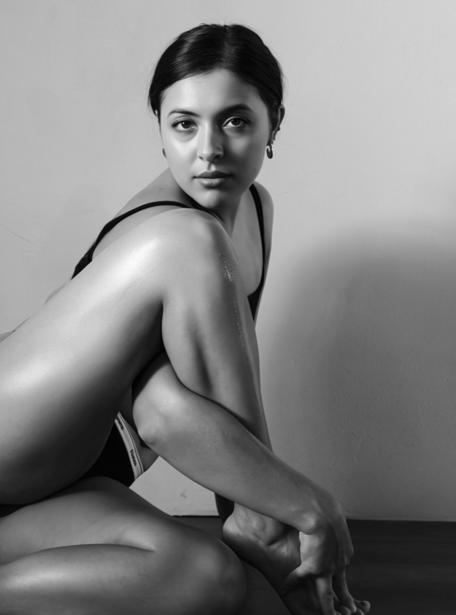 A woman with dark hair and earrings, sitting with her arm resting on her knee, looking at the camera.