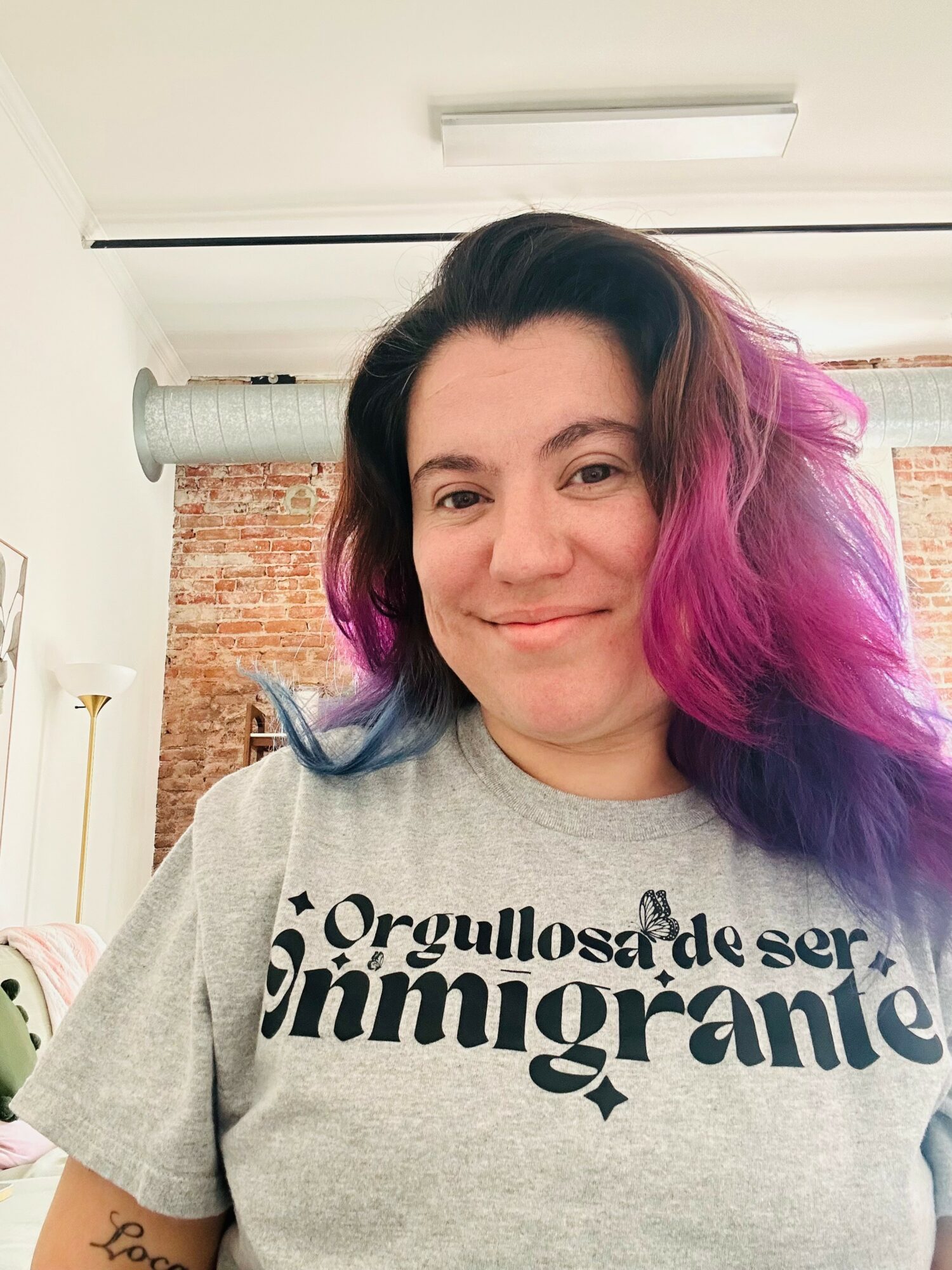 Woman with dark and purple hair smiling indoors, wearing a gray T-shirt with Spanish text.