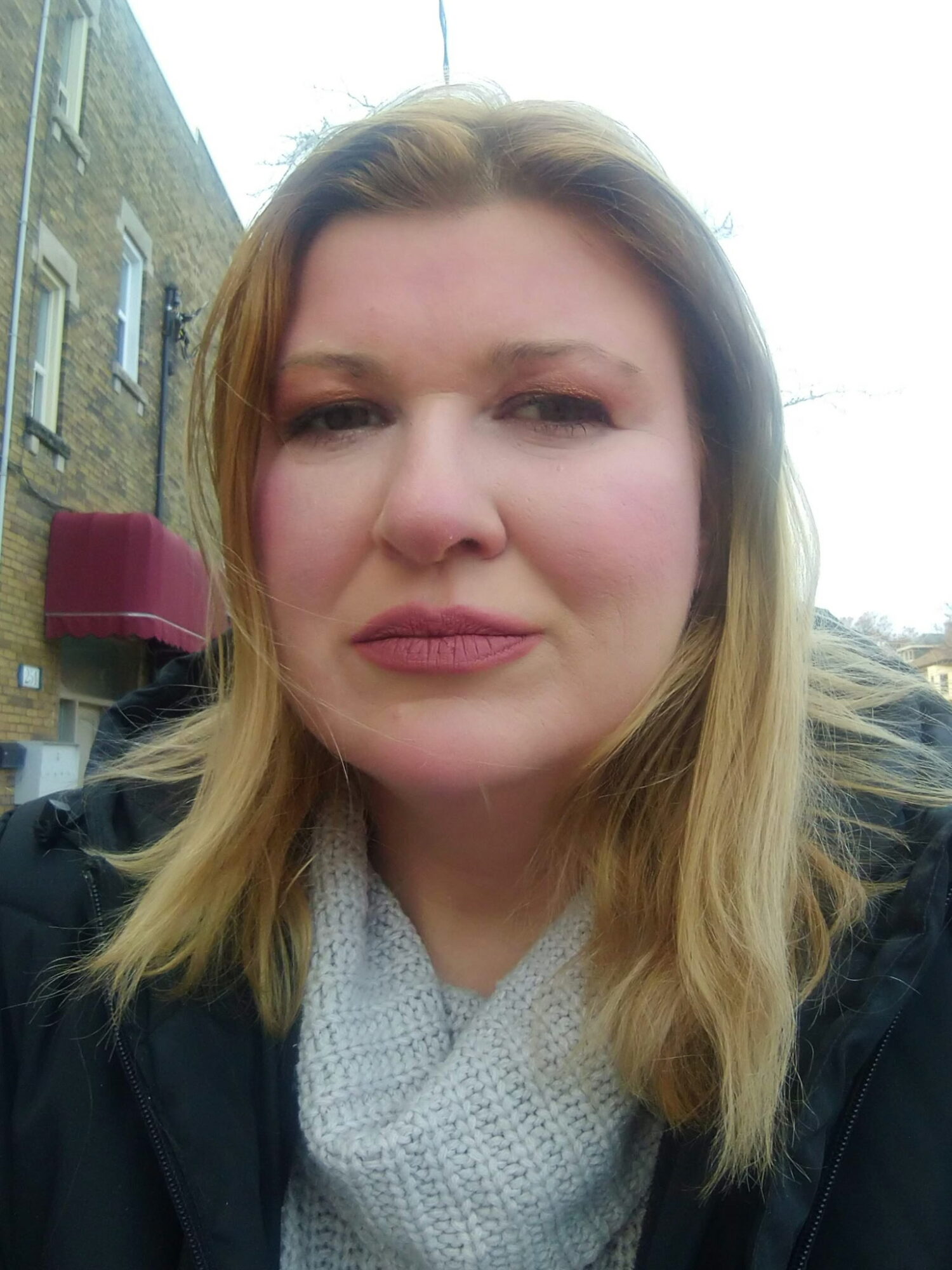 Close-up of a woman with blonde hair outdoors, wearing a black jacket and a light-colored scarf.