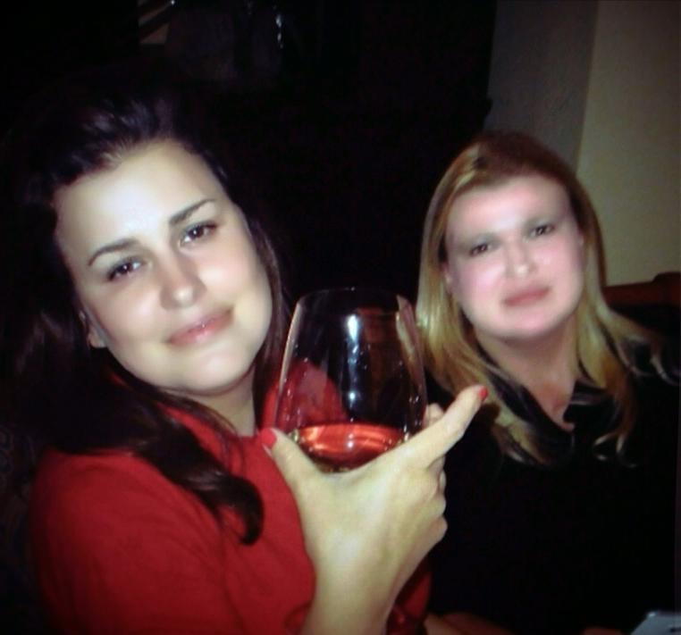 Two women sitting at a table, one holding a glass of red wine, smiling at the camera.