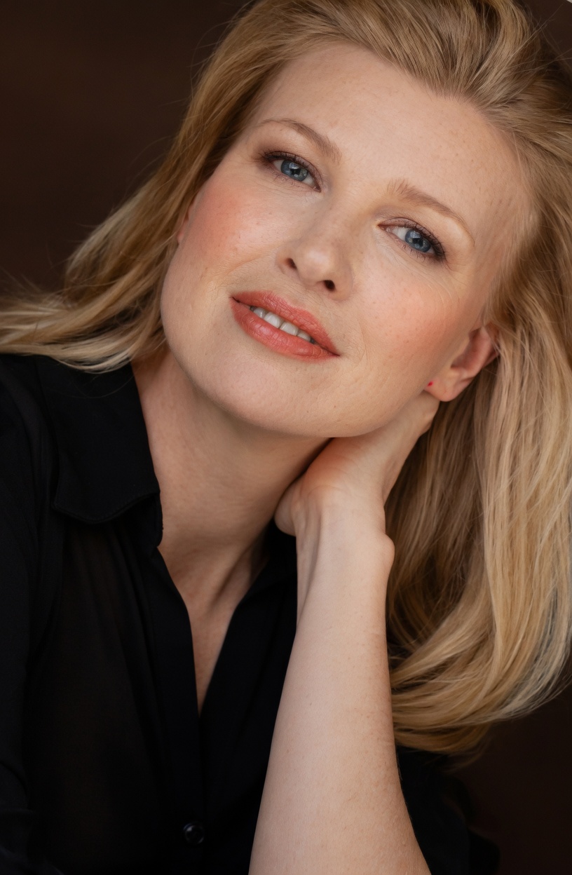 Close-up portrait of a woman with blonde hair, blue eyes, and fair skin, resting her face on her hand, wearing a black top.