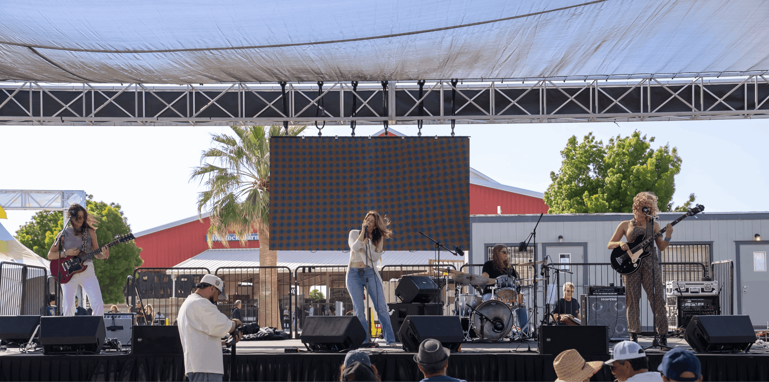 Band performs on an outdoor stage with four musicians, audience in front, trees and buildings in background.