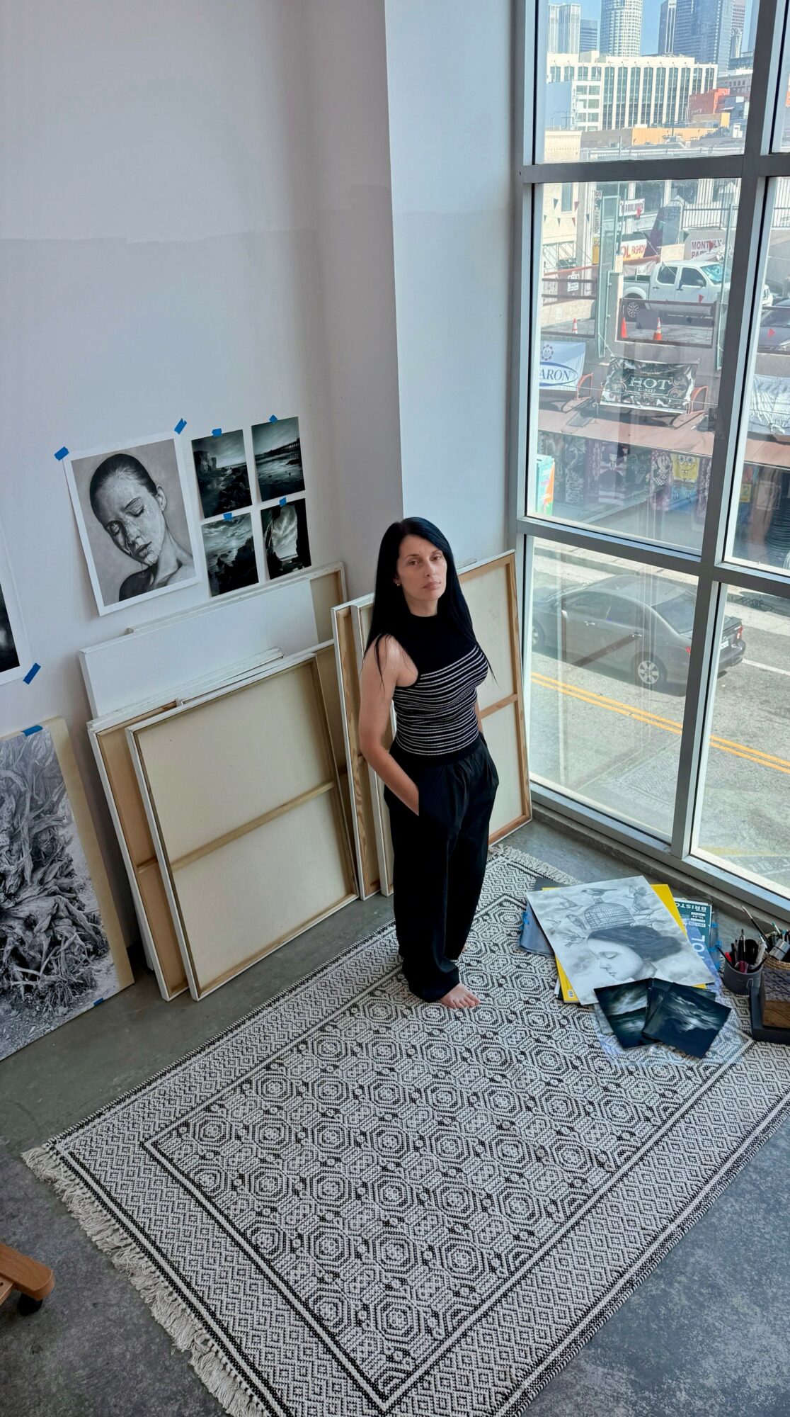 Woman standing in a room with large window, artwork on wall, and a patterned rug on the floor.