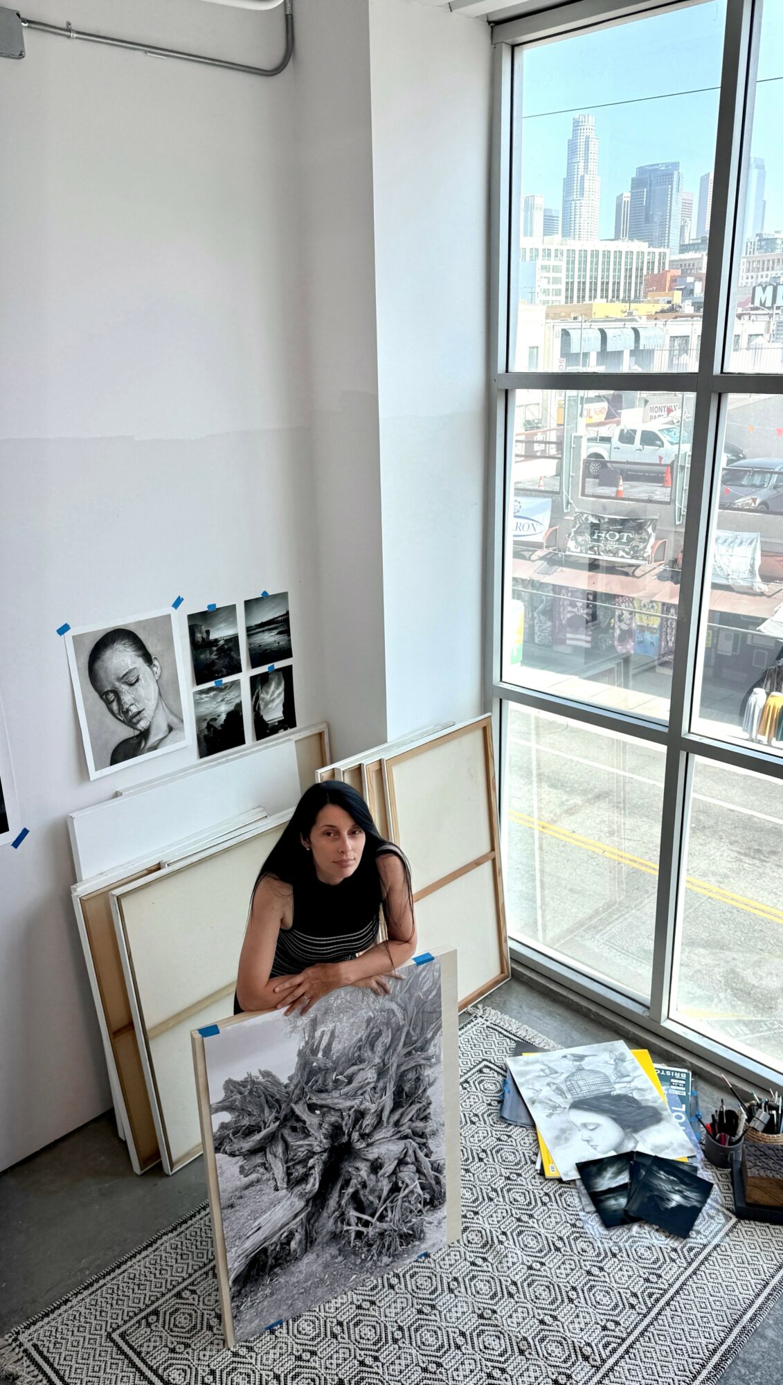 Woman sitting on the floor near large window with city view, surrounded by framed artwork and art supplies, in a bright room.