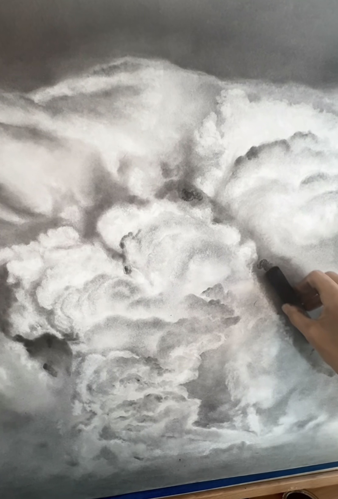 Hand holding a tool drawing detailed clouds in black and white, with various textures and shading.