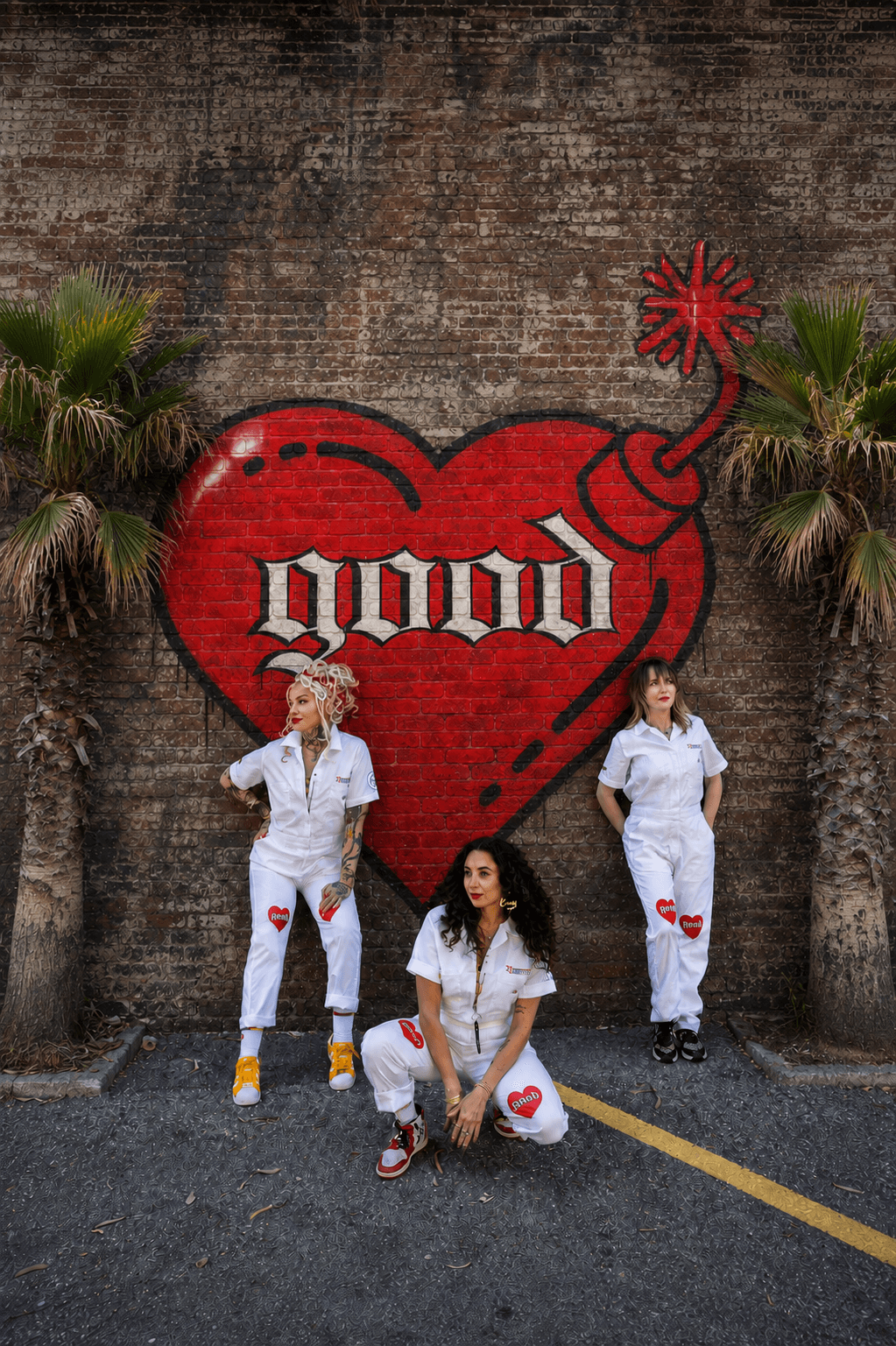Three women in white outfits with red heart patches pose in front of a large red heart mural with the word 'good' and a bomb illustration, against a brick wall.
