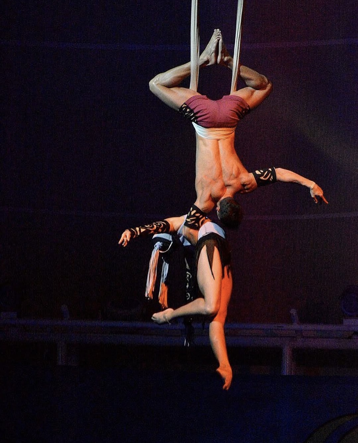 A male aerialist performs an upside-down acrobatic pose on aerial silks, hanging with legs bent and arms extended.