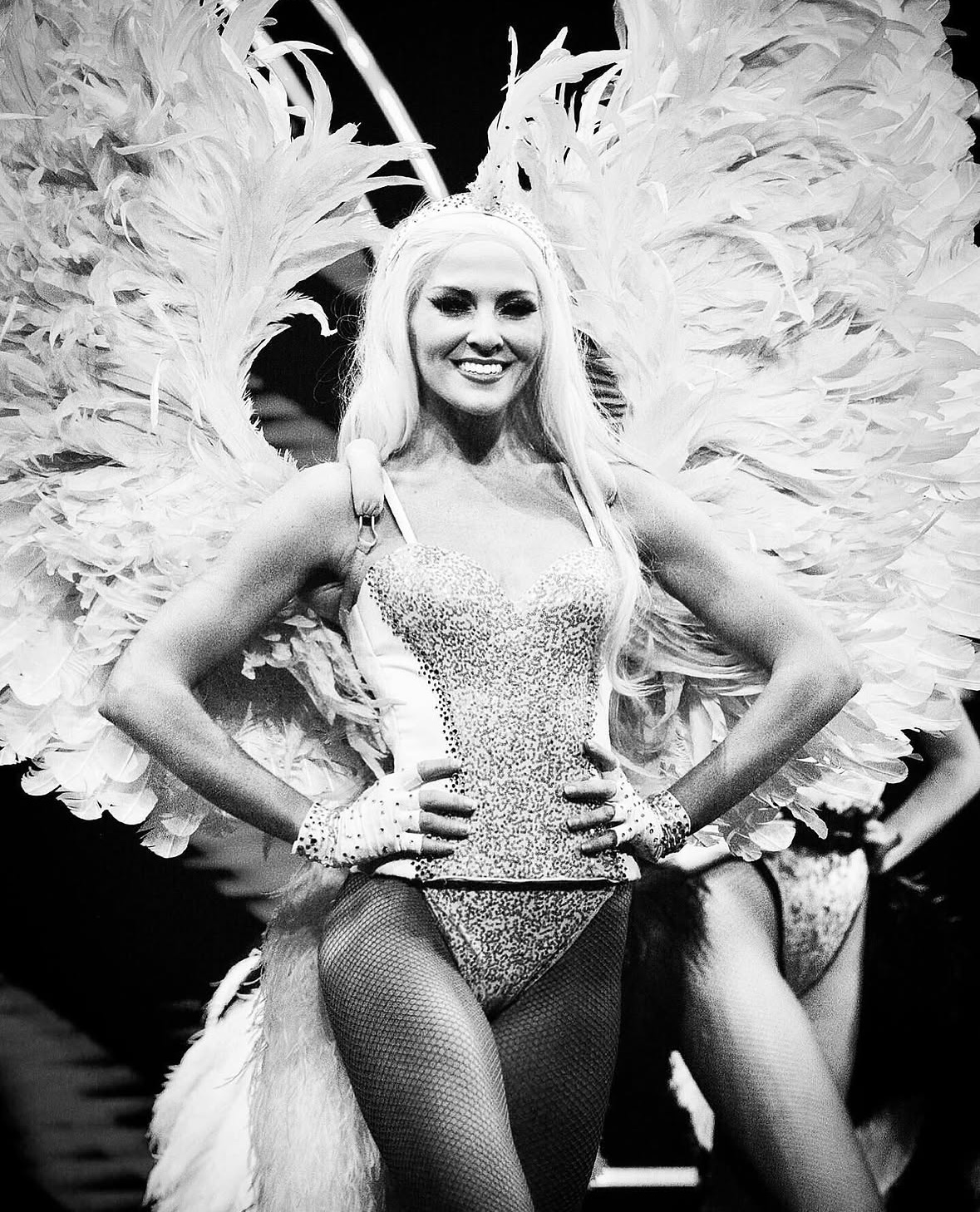 Woman in costume with large feathered wings, smiling, hands on hips, wearing a sparkly outfit and fishnet stockings.
