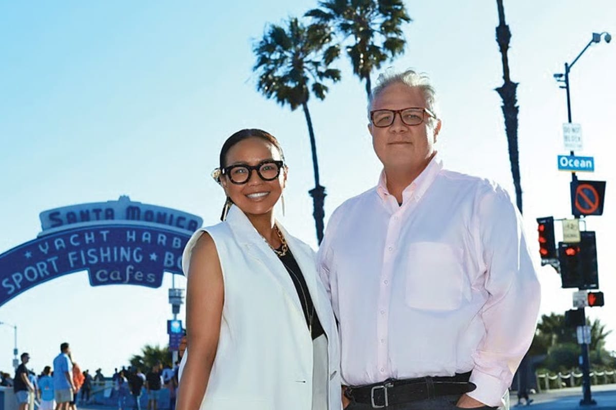 Two people standing outdoors with palm trees and signs in the background, smiling at the camera.